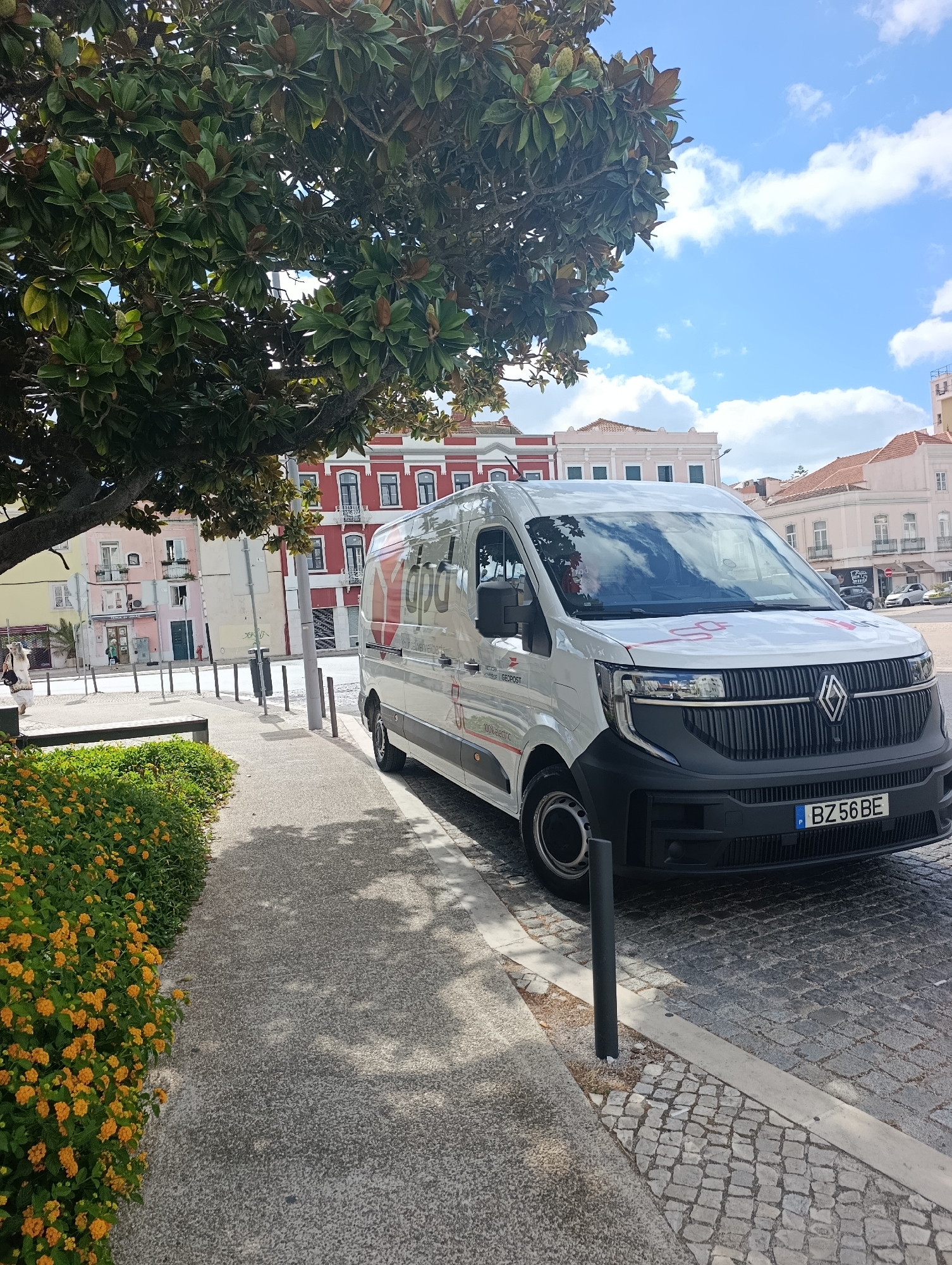 The image captures a bright, sunny day in Setúbal, Portugal, with a white delivery van parked on a cobbled street. The van, which appears to be 100% electric, has the logo "dpa" and "your delivery experts" printed on its side. A license plate with the characters "BZ 56 BE" is visible on the front.
In the foreground, a pathway lined with vibrant orange flowers leads the eye towards the street. A mature tree with large green leaves provides shade, casting dappled shadows on the pathway and the van. Behind the van, buildings with distinct architectural styles are visible, including one with a red facade and white trim, and others painted in pastel colors. The sky is blue with scattered white clouds. A person is walking on the sidewalk in the background. The environment suggests a commercial or residential area in the city.