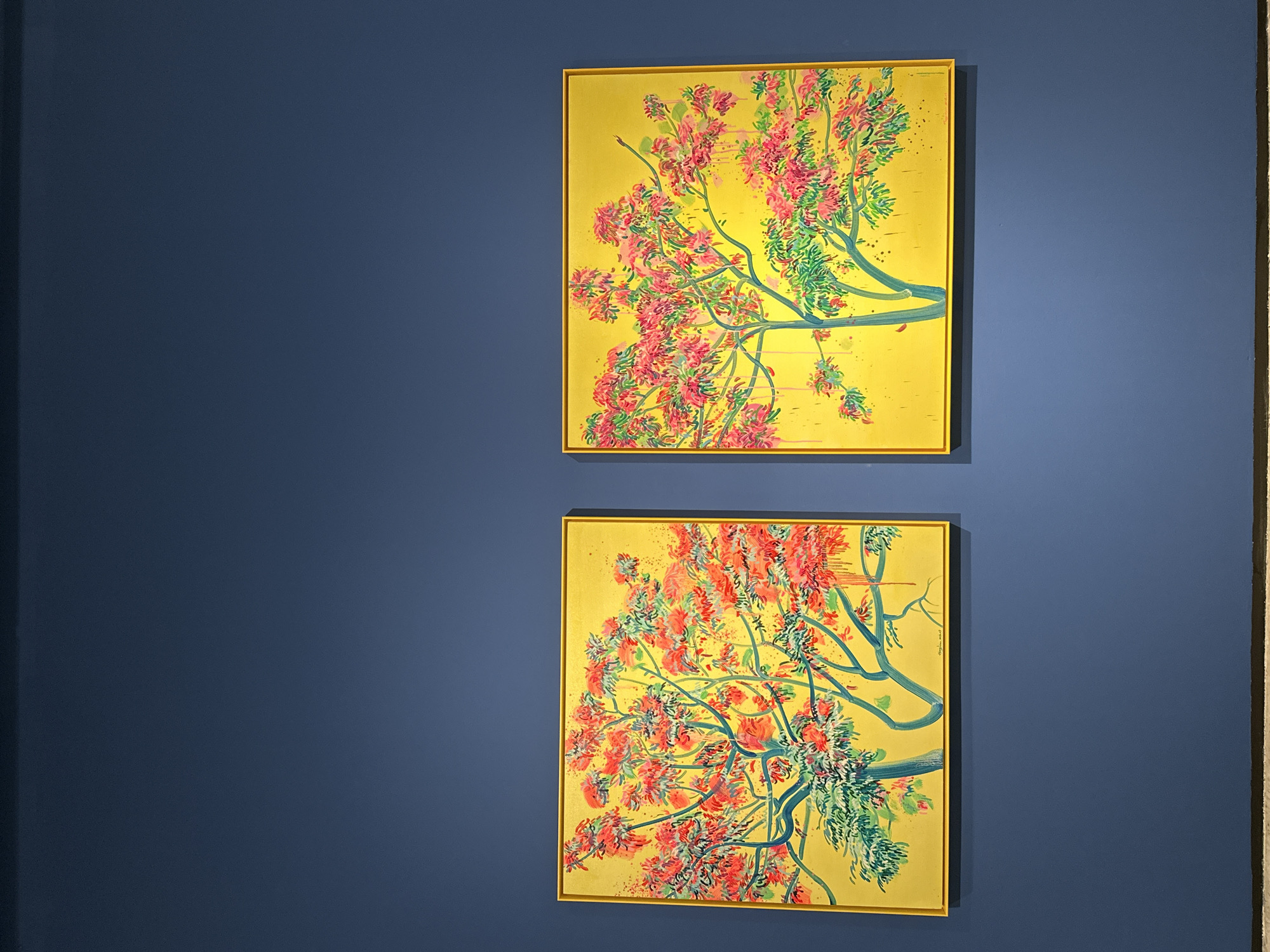 Two framed paintings are hung vertically on a blue wall. The paintings appear to depict a flowering tree or shrub with a yellow background, green branches, and coral-colored flowers. The paintings are identical in style and composition. The wall is smooth and uniformly colored. The scene is likely located inside an art gallery or museum. There are hints of stone or a textured material visible along the right edge of the frame, suggesting a corner of the room or architectural detail. The location in Banan, China, is not visibly discernible from the image.