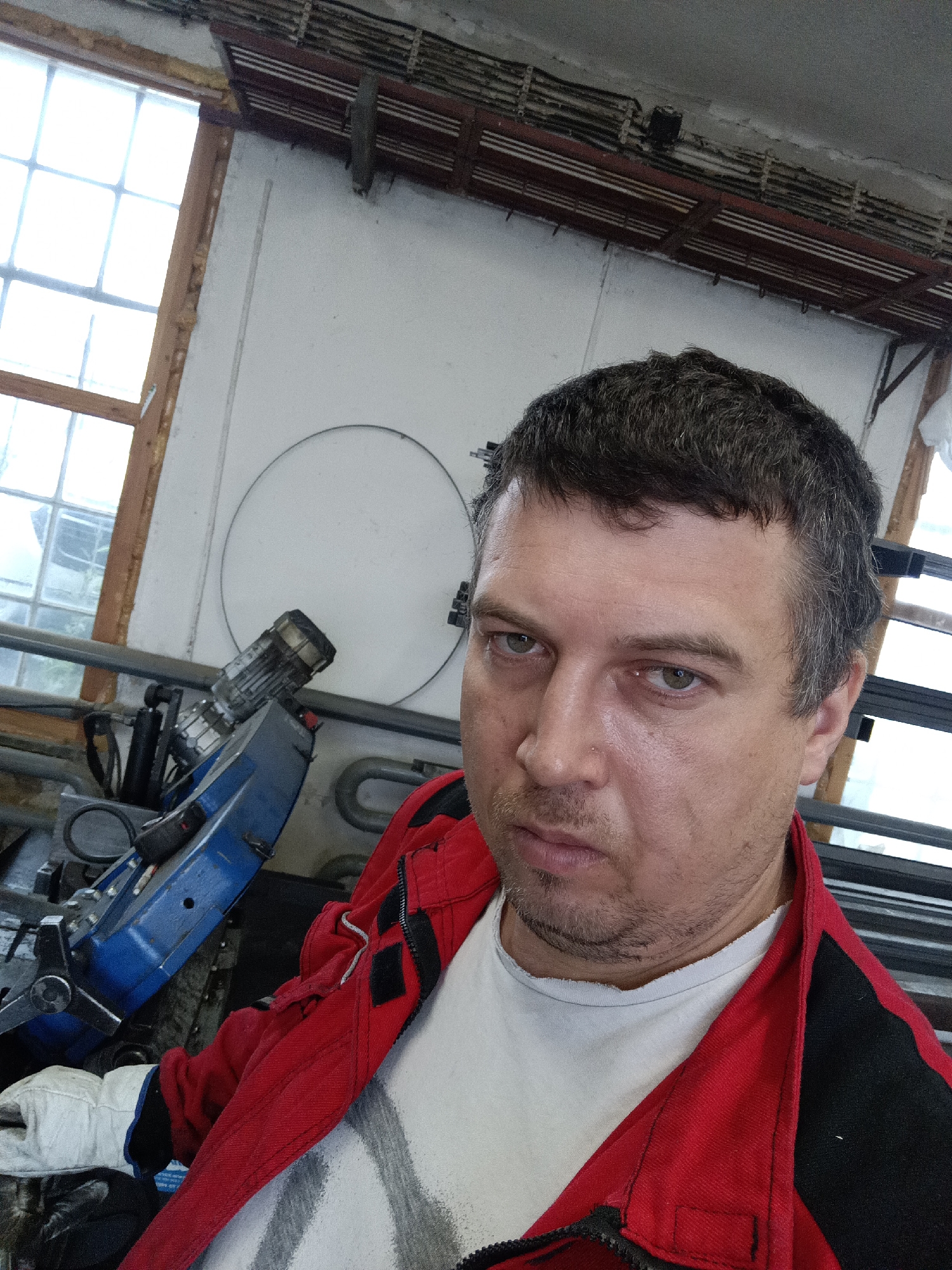 A medium shot photograph shows a man in a red and black work jacket and white t-shirt, looking directly at the camera with a neutral expression. The man has short, dark hair with some graying, and a light beard. He is wearing white work gloves on his left hand.
Behind the man, to his left, is a blue and silver industrial machine with a motor and hydraulic components. To the man's right, there are dark metal structures and in the background, a large window with multiple panes of glass. The environment appears to be a workshop or industrial space, possibly in Dalovy, Czechia, given the context. The walls are white, and there is overhead electrical wiring and lighting fixtures. A large circular object is visible on the white wall behind the machine.