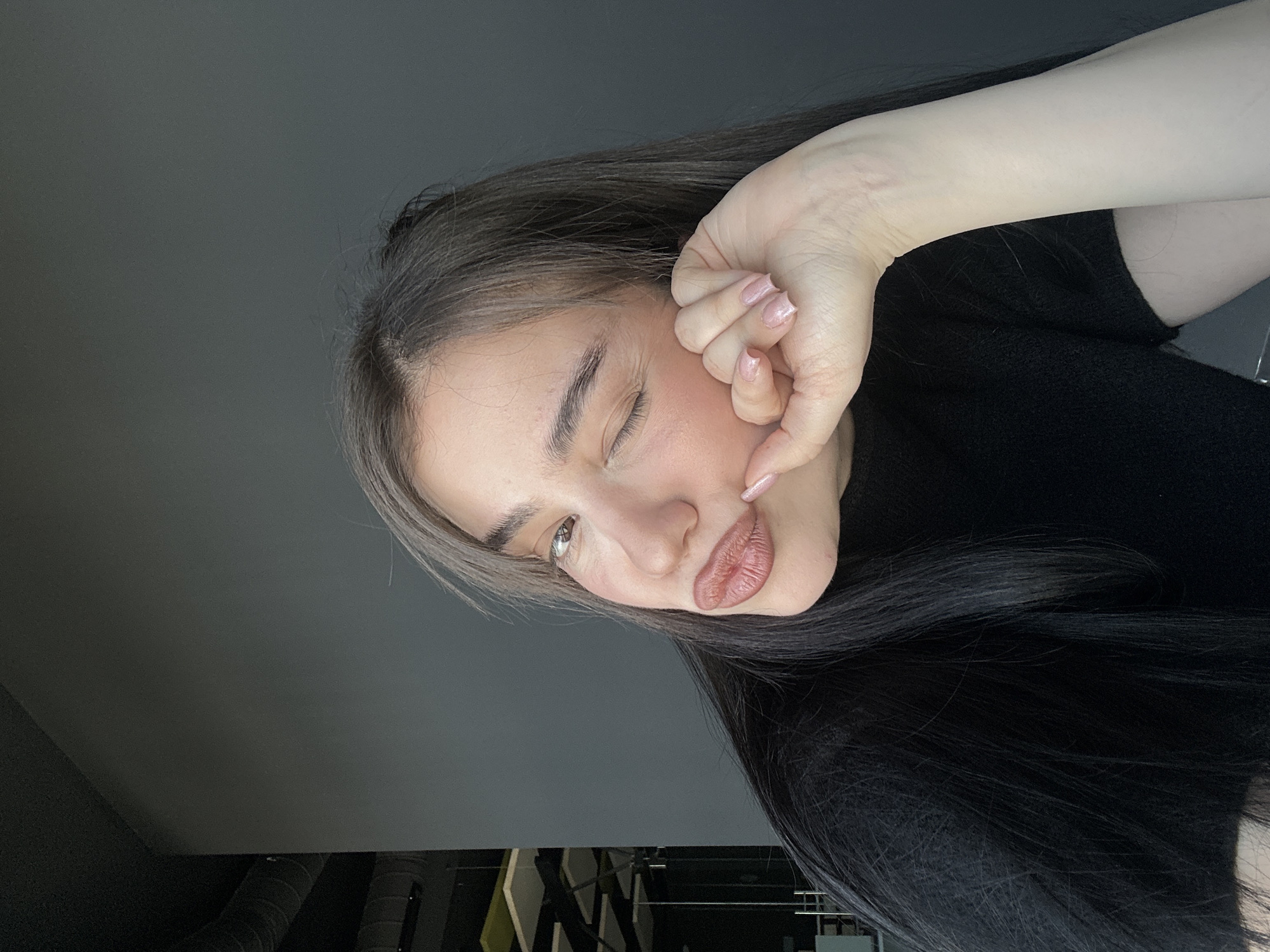 A woman with long, dark hair and light skin is looking directly at the camera with a winking expression. She is holding her hand up to her face, with her fingers touching her cheek. She is wearing a black shirt. The background is a grey wall. The location is unknown; the city of Sofia, Bulgaria, cannot be determined from the image.