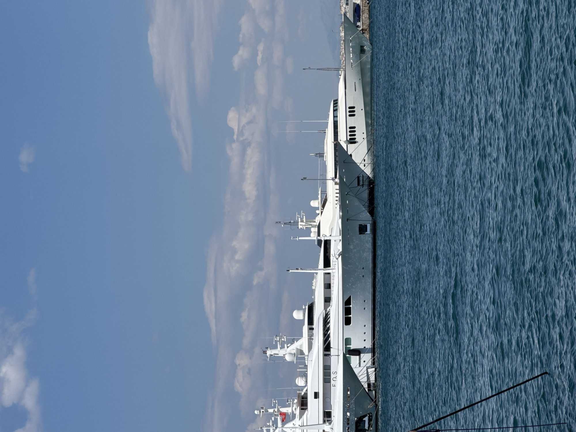 The image shows several large white yachts docked in a harbor. The yachts are lined up side-by-side, with their bows pointing towards the right side of the frame. The water in the foreground is blue and choppy, reflecting the bright sky. In the background, there are buildings and hills visible, suggesting an urban or coastal setting. The sky is blue with scattered white clouds. A red flag is visible on one of the yachts. The yachts appear to be luxurious and well-maintained, indicating they are likely privately owned vessels. There are no people visible in the image. The overall impression is one of a peaceful, sunny day at a marina.
This image was taken in the city of Moschato, Greece, likely at its port facilities or a nearby marina. The presence of multiple large yachts suggests a location frequented by international boaters and tourists. The architectural style of the distant buildings and the natural landscape in the background are consistent with the region. Moschato is a municipality in the Athens metropolitan area, and its port area serves as a gateway to the Saronic Gulf and beyond.