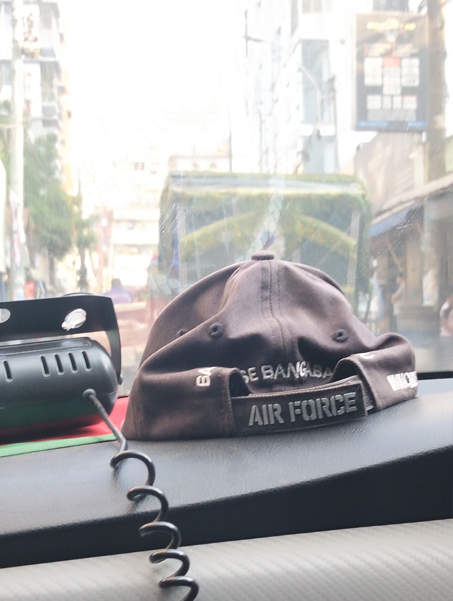The image shows a brown baseball cap on the dashboard of a vehicle. The cap has "BASE BANGLADESH AIR FORCE" embroidered on it. Next to the cap is a black electronic device with a coiled cord. The background is blurred, showing a cityscape with buildings and vehicles, consistent with an urban environment. The presence of "BASE BANGLADESH AIR FORCE" suggests the location is likely Dhaka, Bangladesh.