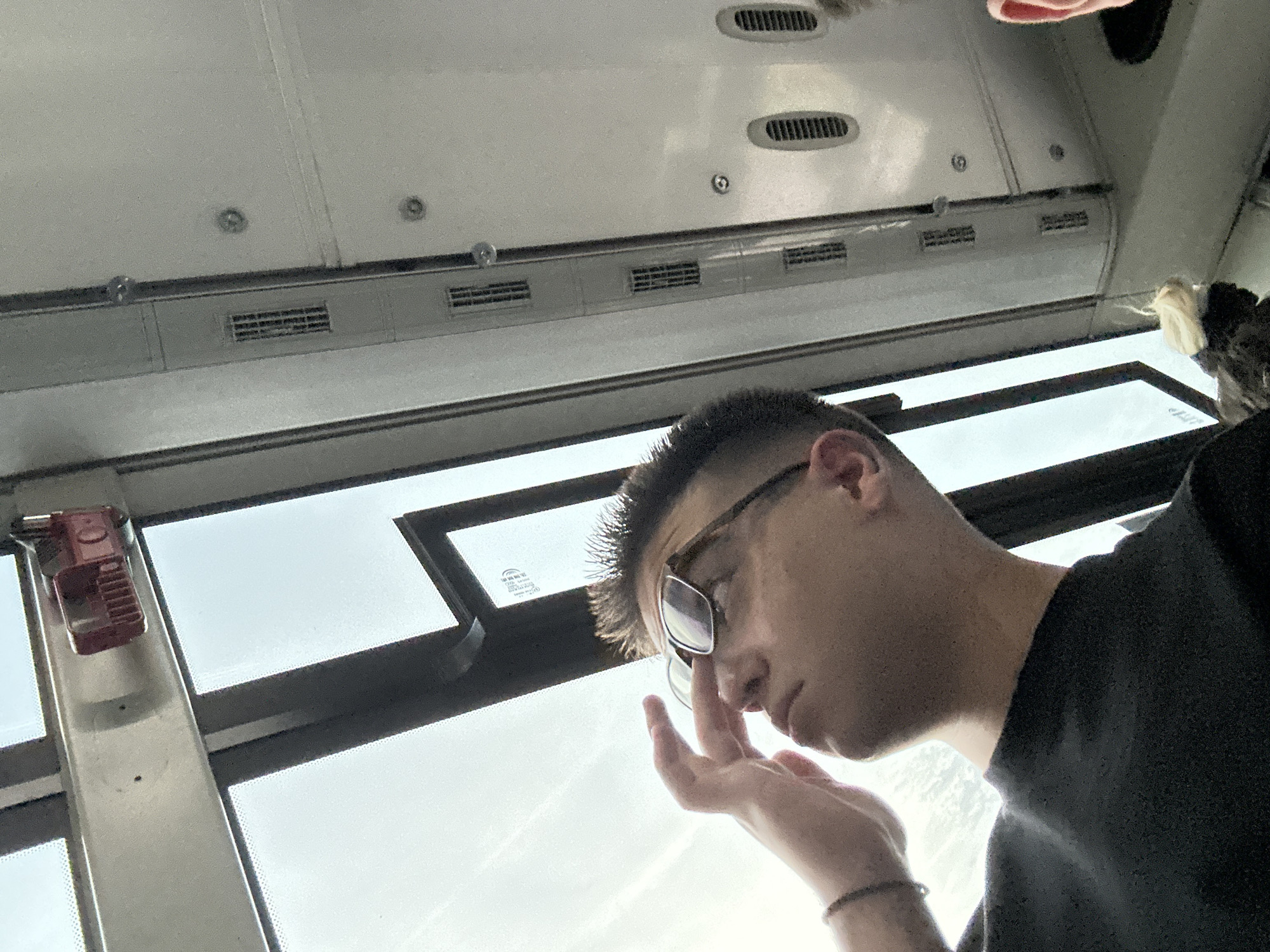 A close-up, upward-angled shot shows a young man with short hair and glasses looking up towards a bright sky through a window. He is wearing a black t-shirt and has his right hand near his face, touching his nose. The interior of a vehicle, possibly a bus or train, is visible with metal panels and vents along the ceiling. A red emergency pull handle is mounted on the window frame above him. The lighting suggests it is daytime.