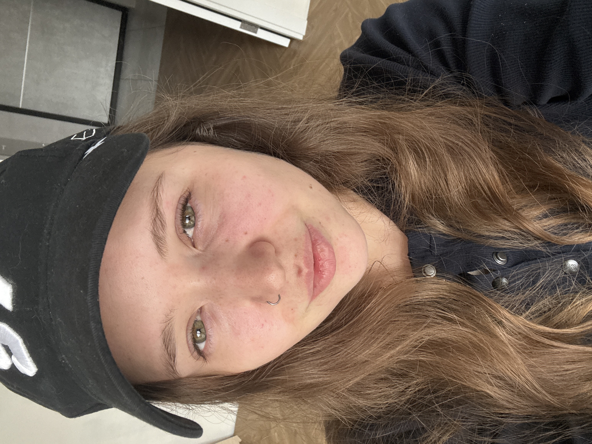 A selfie captures a young woman with light skin, freckles, and a nose ring. She has light brown hair and green eyes. She wears a black cap with white lettering on the side. Her clothing appears to be a dark blue or black button-down shirt. The background shows a white cabinet and a tiled wall. No location in Izhevsk, Russia, can be determined from the given information.