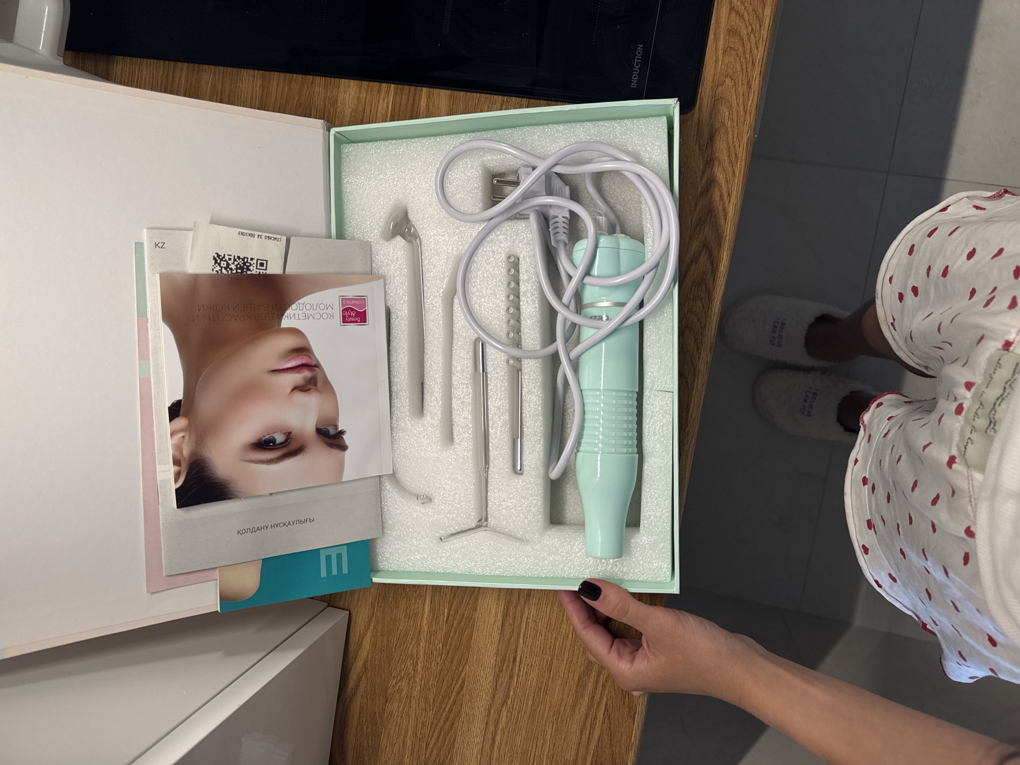 The image depicts a high-angle overhead view of a person's upper body and lower legs, situated in a domestic setting within the city of Chaikovsky, Russia. A pair of hands, one with black-painted fingernails, holds open a light-green rectangular box containing a beauty or medical device. The box's interior features white foam padding meticulously shaped to secure a light teal-colored handheld electronic device, characterized by a ribbed grip and an attached coiled white power cord terminating in a two-prong European-style plug. Adjacent to the main device, multiple clear glass or metallic wand-like attachments of varying shapes are nestled within the foam.
Overlapping the upper section of the box are several informational booklets and papers. One prominent booklet displays a close-up image of a woman's face and features text in Russian and Kazakh, including "КОСМЕТИКА ДЛЯ КРАСОТЫ И МОЛОДОСТИ ВАШЕЙ КОЖИ" (Cosmetics for the beauty and youth of your skin) and "КОЛЛАГЕНУ НУСКАУЛЫҒЫ" (Collagen Advice). A "Beauty Style Cosmetics" logo is also visible. Another small paper piece displays a QR code and the Russian phrase "СПАСИБО ЗА ПОКУПКУ" (Thank you for your purchase), with "KZ" printed above it.
The box rests on a light brown wooden countertop or table. To the left, a white cabinet or wall surface is visible. To the right, a black appliance, likely an induction stovetop, shows the word "INDUCTION" printed on its surface. Below the countertop, the person's legs are visible, clad in white shorts or pajama bottoms adorned with small red heart patterns. The person wears white, fluffy slippers, one of which partially displays the text "I BELIEVE I CAN FLY." The floor beneath is composed of light gray tiles.