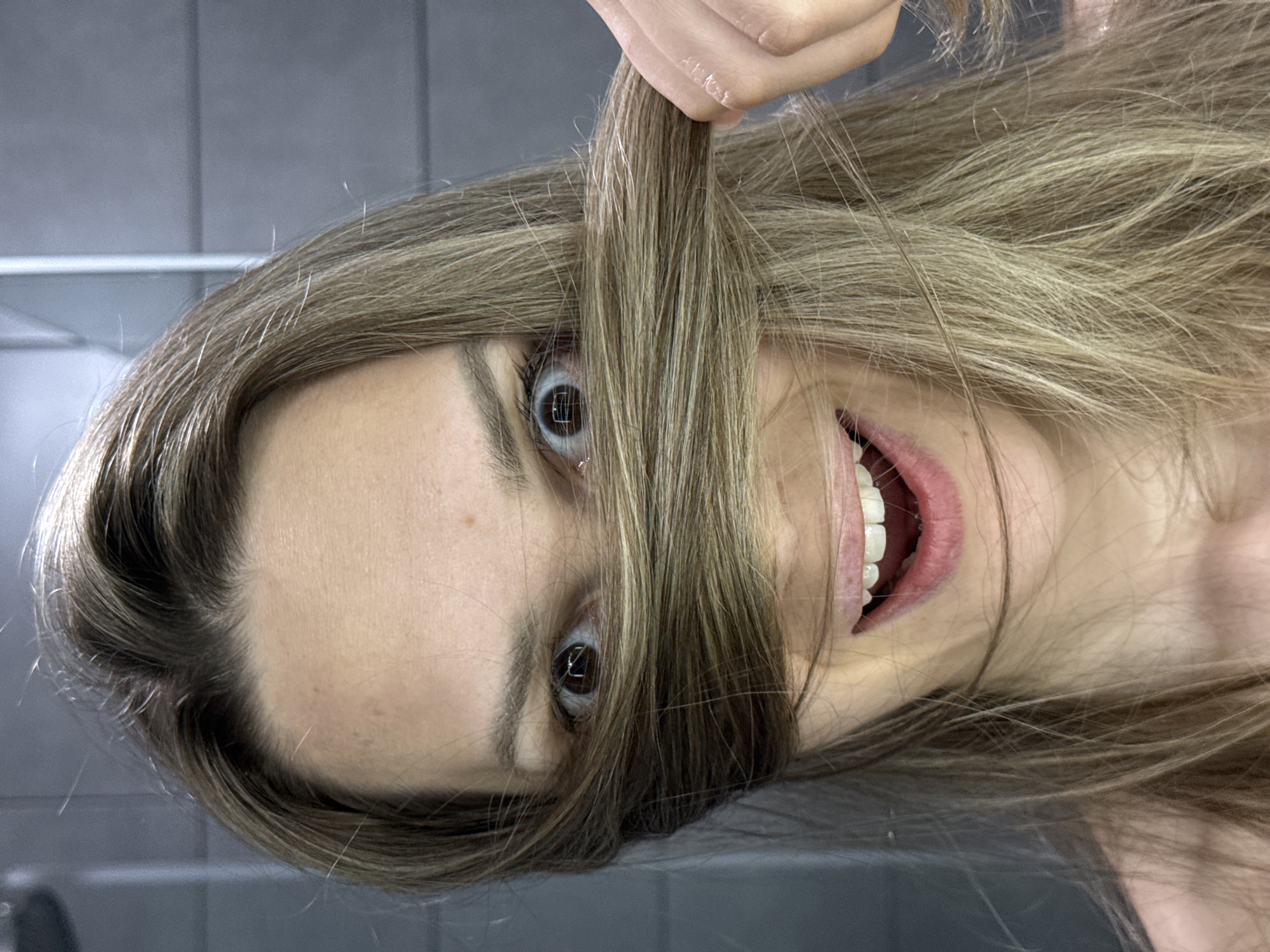 A woman with brown hair and brown eyes is upside down in front of a gray tiled wall. She is holding a strand of her hair in front of her face, obscuring her vision. She is smiling, revealing her teeth. The location is not identified as Chaikovsky, Russia.
