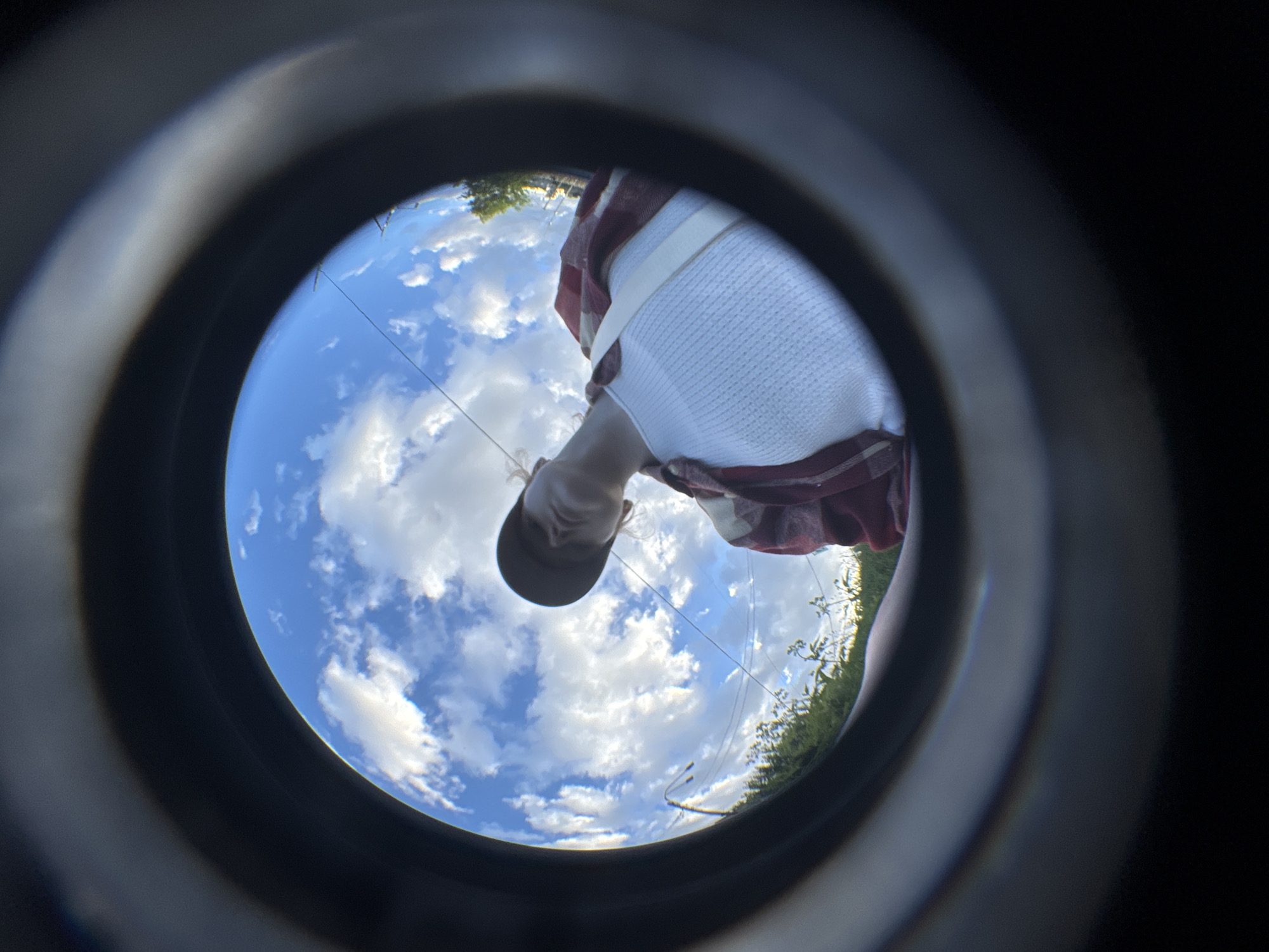 A low-angle, wide-angle fisheye lens perspective captures a person from below, looking up towards a bright blue sky filled with white, fluffy clouds. The subject, wearing a red and white plaid shirt over a white textured tank top and a dark baseball cap, is positioned in the upper right quadrant of the frame. Their head is tilted back, and their face is partially obscured by the cap, but their gaze appears directed upwards. In the background, the sky is dominated by the clouds, with a hint of greenery and trees along the horizon line, suggesting an outdoor setting. The overall impression is one of a person observing the sky in what appears to be a serene, open environment. The image does not provide any specific details or visual cues that would allow for the identification of the location as Saint Petersburg, Russia, or any other city.