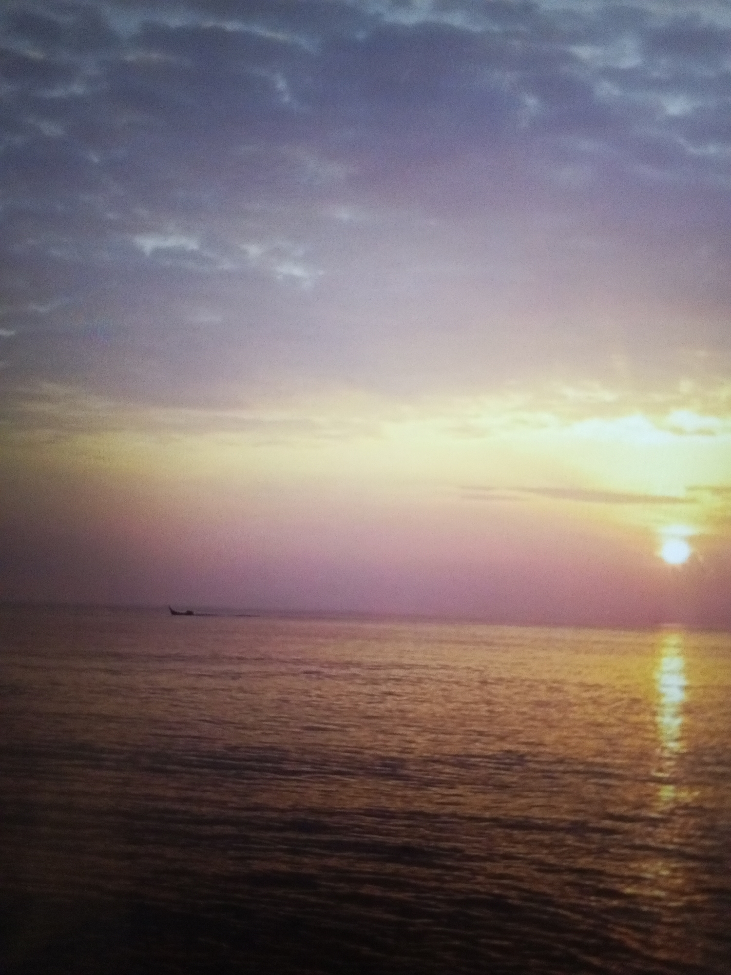 The image captures a sunset over the ocean in Lhokseumawe, Indonesia. The sky transitions from deep purple and gray clouds at the top to bands of lighter pinks, yellows, and oranges near the horizon. The sun, a small, bright orb, sits on the horizon, casting a golden reflection on the water. A small boat is visible in the distance, towards the left side of the frame. The water is calm, with gentle waves reflecting the colors of the sky.