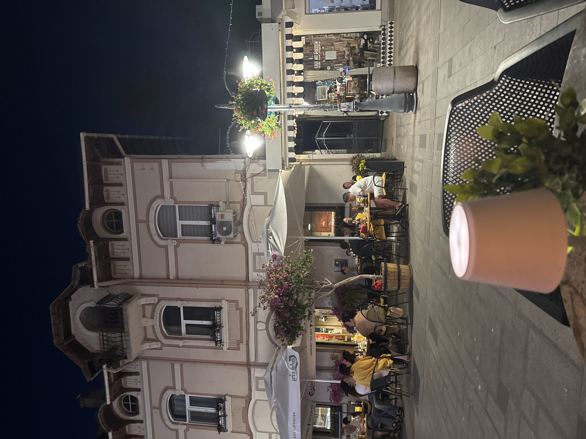The image captures an outdoor dining scene at night on a European-style street, likely in Burgas, Bulgaria, given the architectural style. In the foreground, a metal table and chair are visible, with a pink lamp shade and a small plant in focus. Beyond this, several outdoor seating areas are occupied by patrons enjoying dinner. The establishments appear to be restaurants or cafes, with umbrellas providing shade. A multi-story building with classical architectural elements, including arched windows and a balcony, forms the backdrop. Streetlights and hanging flower baskets illuminate the scene. A shop with the sign "GIGI ART & DESIGN" is visible to the right, showcasing its wares through a large glass window. People are casually dressed, engaged in conversation and dining. The overall atmosphere suggests a lively evening at a restaurant or cafe.