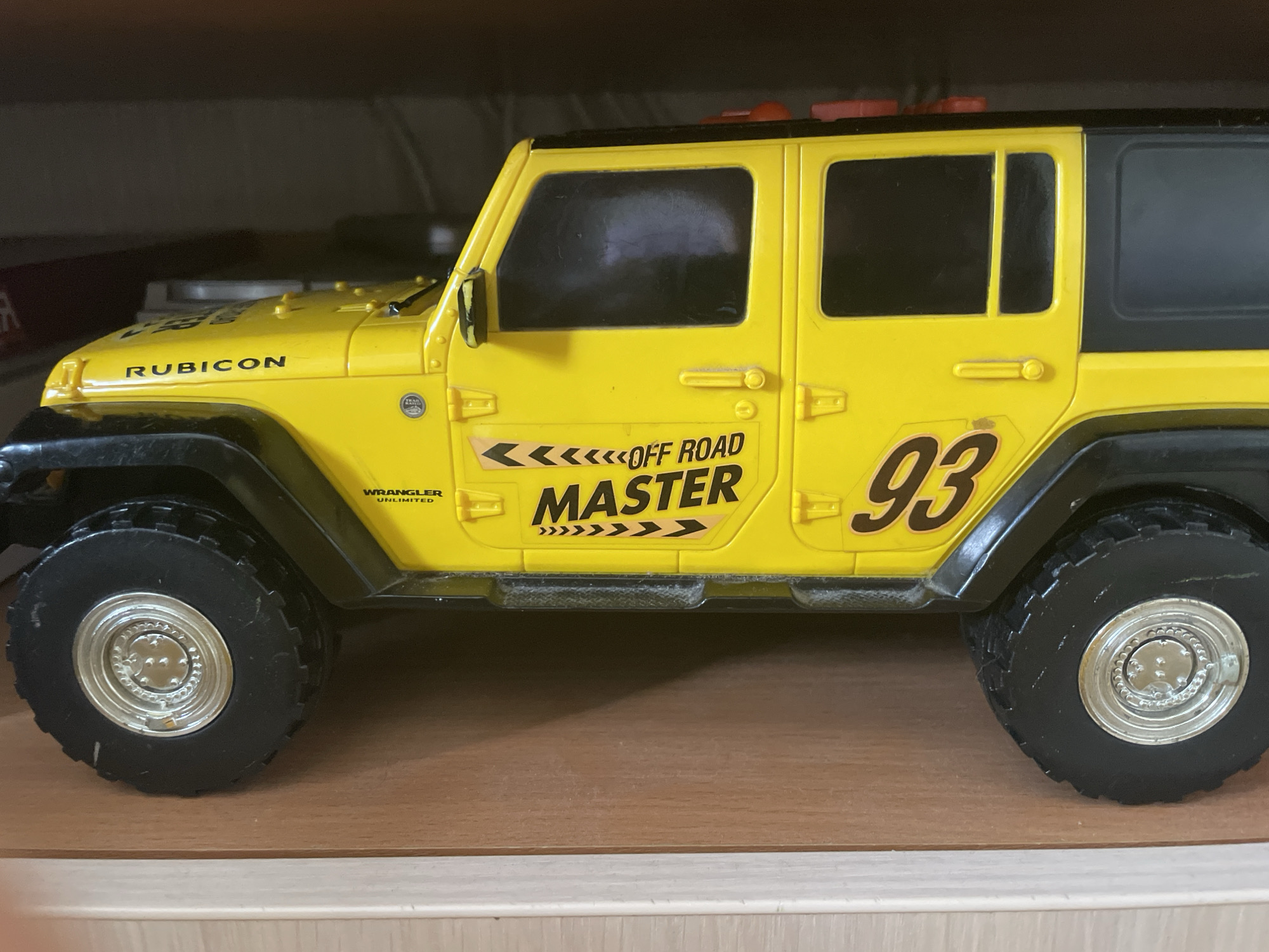 A yellow toy Jeep Wrangler Rubicon Unlimited is on display. The Jeep features black accents on the tires, fenders, and roof. The side of the Jeep has "OFF ROAD MASTER" graphics and the number "93". The toy is positioned on a wooden shelf with a dark background.