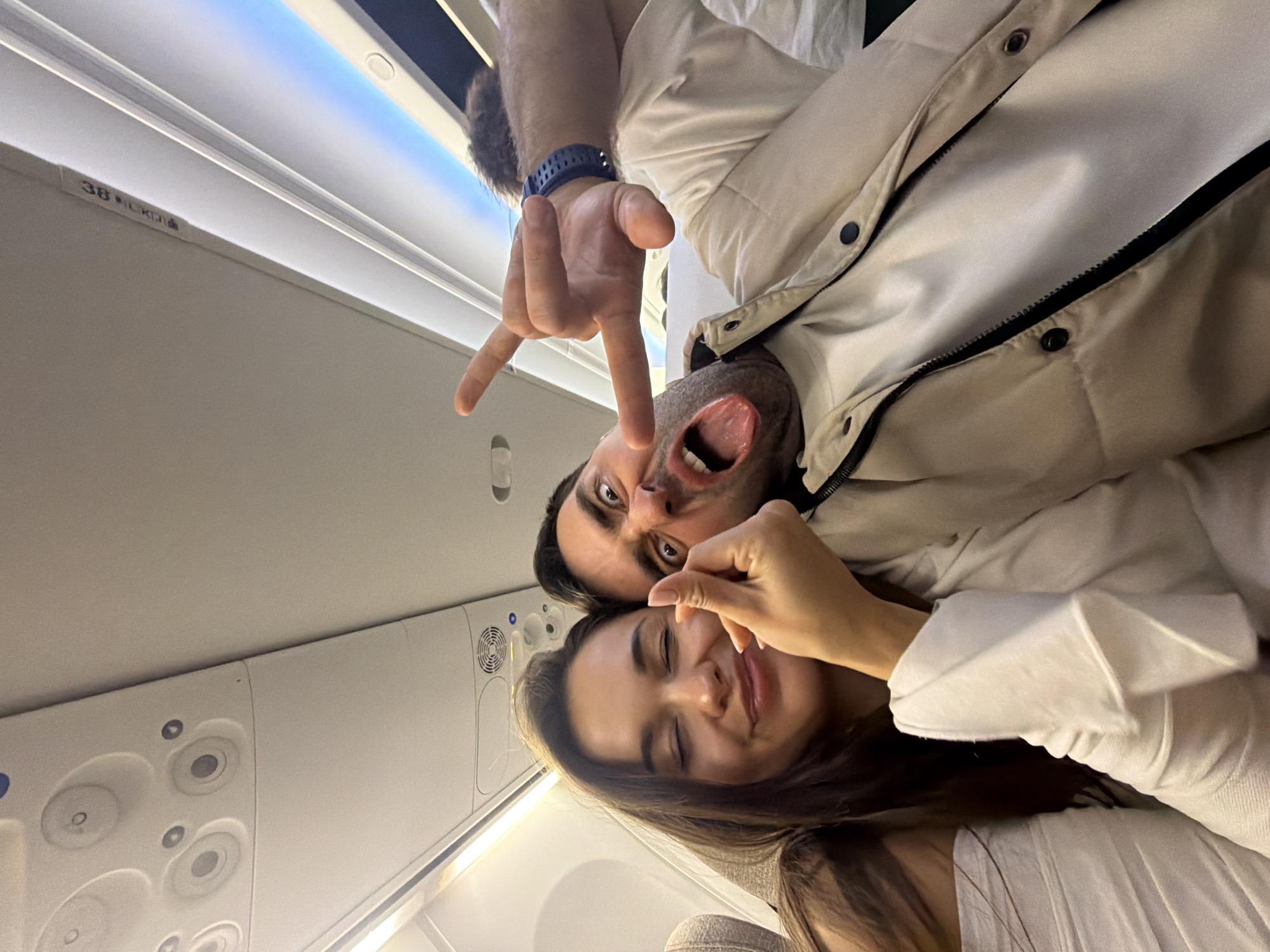 A selfie-style photo taken inside an airplane depicts two people. A man is at the top of the frame, making a playful face with his tongue out and eyes wide open. His hand is raised, forming a "rock on" hand gesture. He is wearing a white jacket, and a blue smartwatch is visible on his wrist.
Below him is a woman with long, dark hair, smiling and squinting her eyes. Her hand is near her face, as if she is pinching her nose. She is wearing a white shirt.
The background reveals the interior of an airplane, showing overhead compartments, lights, and air vents. Seat number "38" is visible, and this scene does not depict any specific details that would indicate a location in Zhangjiajie, China.