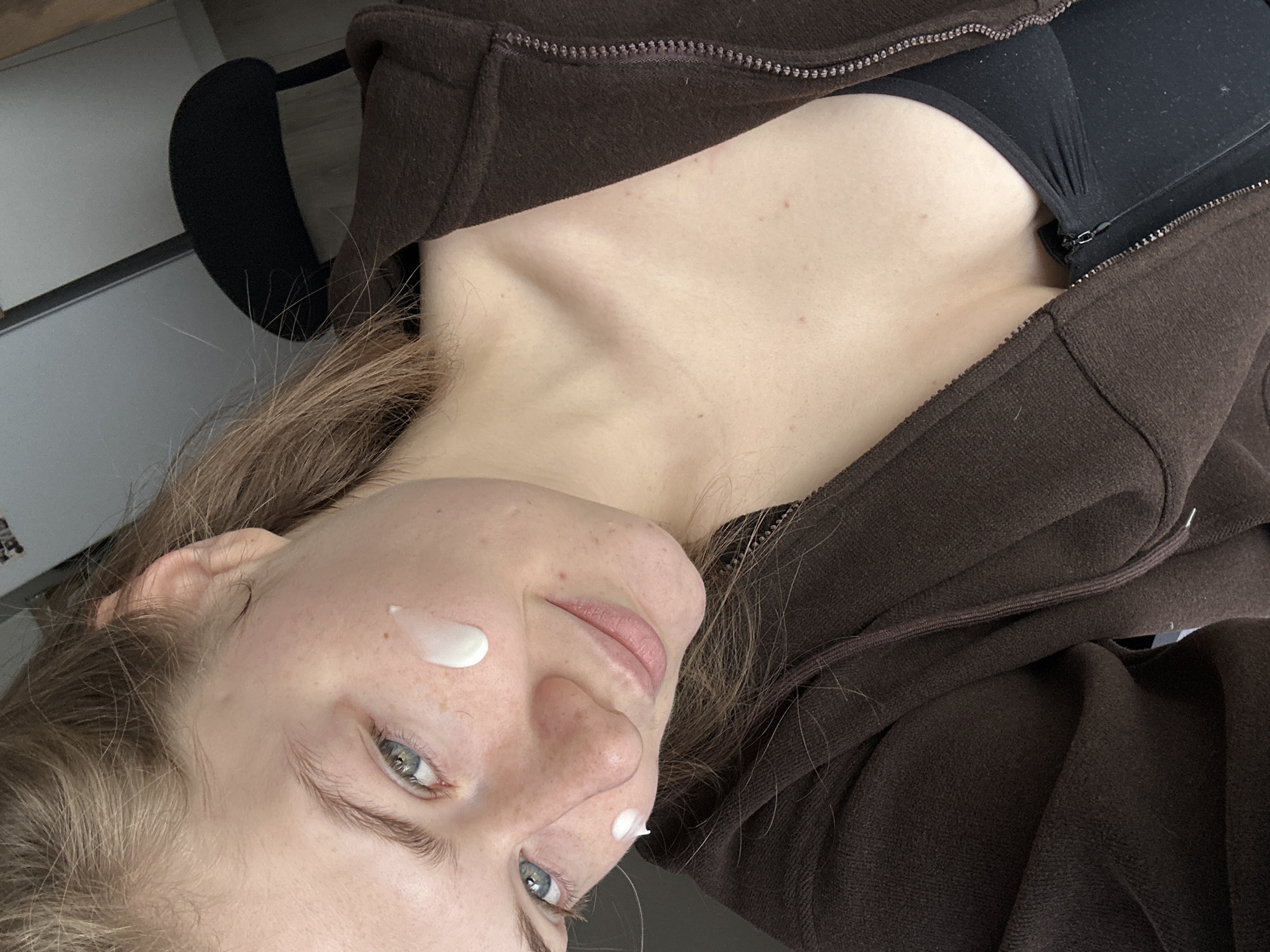 A close-up, angled shot shows a person with fair skin, blonde hair, and blue eyes. The person has a few dots of white cream on their face. They are wearing a brown zip-up hoodie over a black top. A black object is partially visible behind their head. The location and any reference to the city of Kazan, Russia, cannot be determined from the image.