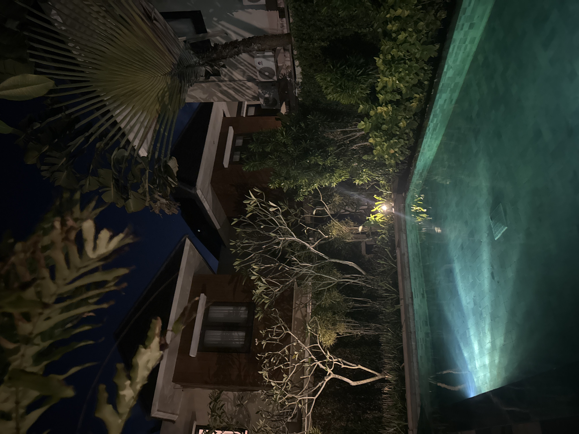 The image depicts a nighttime scene in Kuta Selatan, Indonesia, featuring a building complex adjacent to a pool. The buildings are multi-storied, with exterior walls appearing to be painted in neutral tones and dark-framed windows. Lush tropical foliage, including palm trees and various shrubs, flanks the buildings and pool area. The pool itself is illuminated, reflecting light and creating a rippled pattern on the water's surface. Air conditioning units are visible on the exterior of one of the buildings. The lighting is dim, suggestive of a low-lit or nocturnal setting.