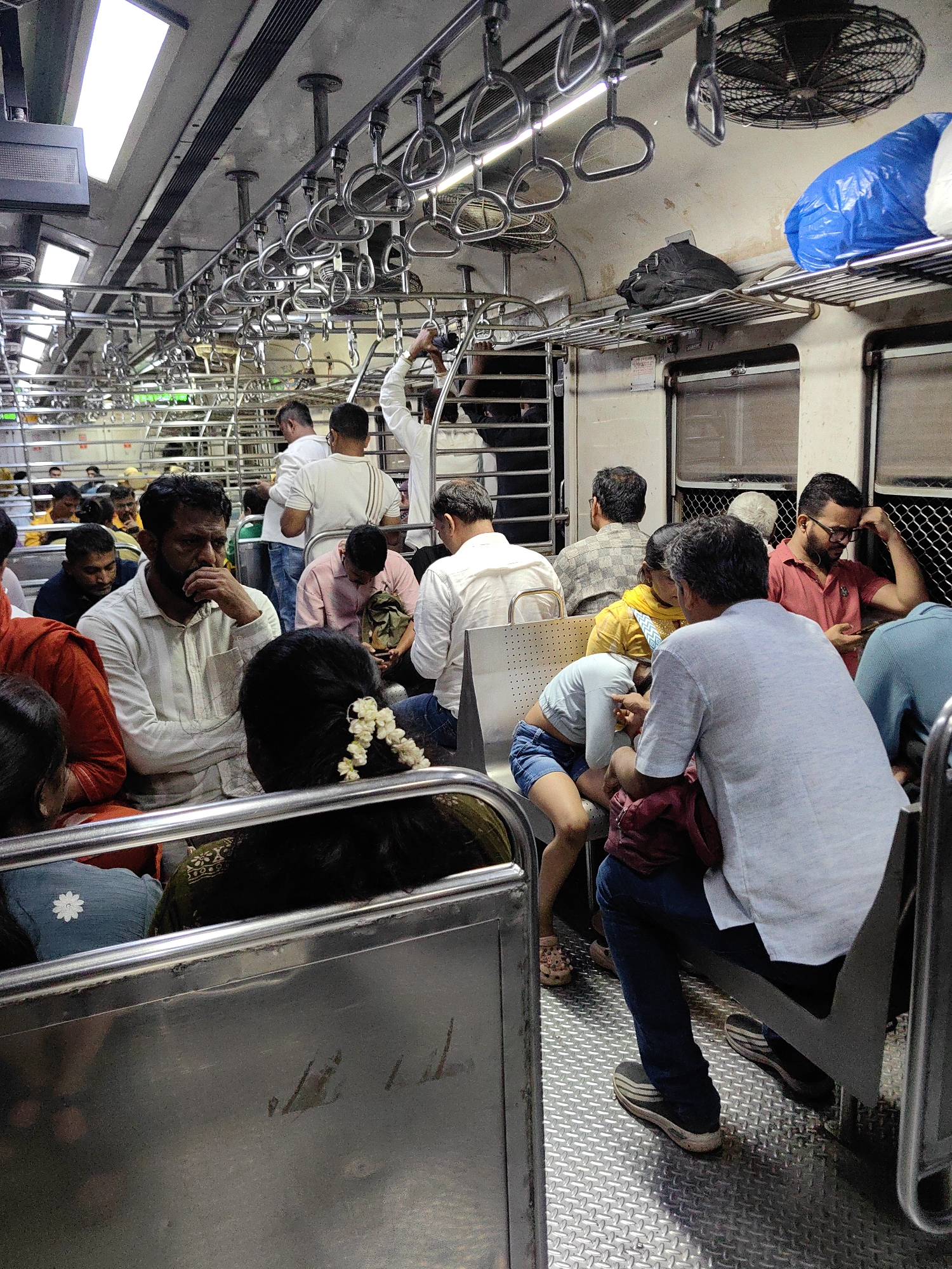 The image depicts the interior of a crowded local train carriage in Mumbai, India. Numerous passengers are seated and standing in close proximity, suggesting a peak travel time. The carriage is equipped with metal handrails and luggage racks, typical of Mumbai's suburban railway system. Passengers are dressed in casual attire, and some are carrying bags. The lighting appears to be artificial, contributing to the somewhat dim and enclosed atmosphere of the train. The floor is metallic, likely stainless steel, with a diamond plate pattern for grip. The overall scene conveys a sense of daily commuter life in a bustling metropolis.