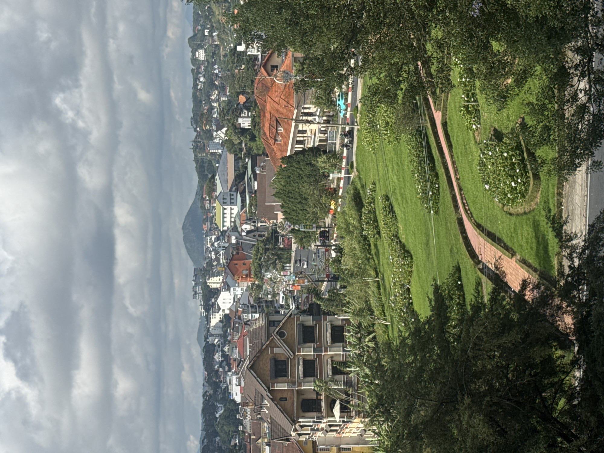 This image captures a panoramic view of a city with a hilly landscape, characteristic of Da Lat, Vietnam. In the foreground, a well-manicured green lawn is punctuated by a winding brick pathway, leading through clusters of white flowers and neatly trimmed hedges.
Dominating the mid-ground are several buildings. On the left, a multi-story house with a distinct architectural style, featuring brown and beige facades and dark-framed windows, stands prominently. To its right, a street with moderate traffic is visible, with cars and motorbikes navigating the road. Further into the scene, more buildings with red-tiled roofs and varied architectural designs are scattered across the slopes. One building with an orange facade and a prominent red roof stands out.
The background reveals a dense urban landscape stretching across undulating hills, dotted with numerous houses and some taller buildings. In the far distance, a mountain range is faintly visible under a cloudy sky. The overall impression is that of a vibrant city nestled within a verdant, mountainous terrain. The lighting suggests it is daytime, possibly late morning or early afternoon, given the shadows cast by the trees and buildings.