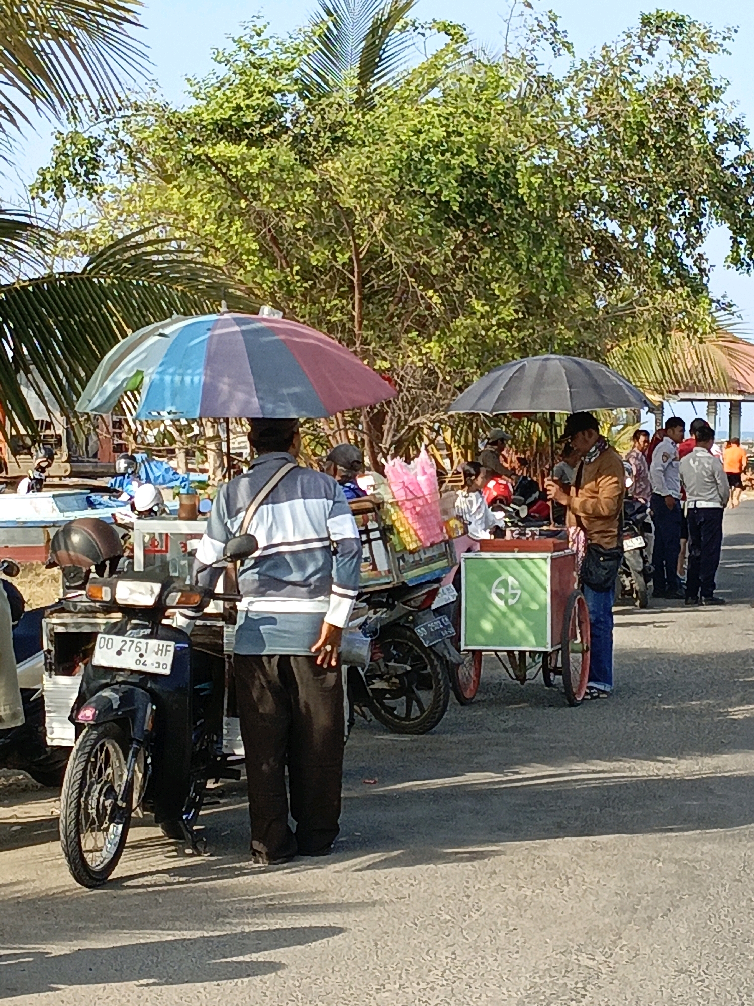 In Bulukumba, Indonesia, a street scene unfolds under a bright, sunny sky, with palm trees and lush green foliage forming a backdrop. The foreground features a road where several individuals are gathered around food carts and motorcycles.
A man in a striped long-sleeved shirt and dark pants stands near a motorcycle with a license plate reading "DD 2761 HF". He appears to be observing the activity around a food cart. The cart, adorned with a green panel and a circular symbol, is sheltered by a black umbrella, under which a man in a brown jacket and blue jeans is interacting with the cart.
Another food cart, larger and equipped with a colorful umbrella featuring red, blue, and purple sections, is also present. This cart is laden with packaged goods, some in pink plastic. Several people, including a woman in a white top, are clustered around this cart.
Further down the road, a group of men in uniform, possibly security or police personnel, are standing together. One man wears a white shirt with dark pants and a badge, another wears a light blue shirt and dark pants, and a third is in a white shirt with dark pants. They are positioned near motorcycles.
The overall impression is a bustling street market or a roadside food stall area, typical of a coastal or tropical urban environment. The presence of numerous motorcycles suggests a common mode of transportation in the area. The sunlight casts distinct shadows on the asphalt, indicating a clear day.