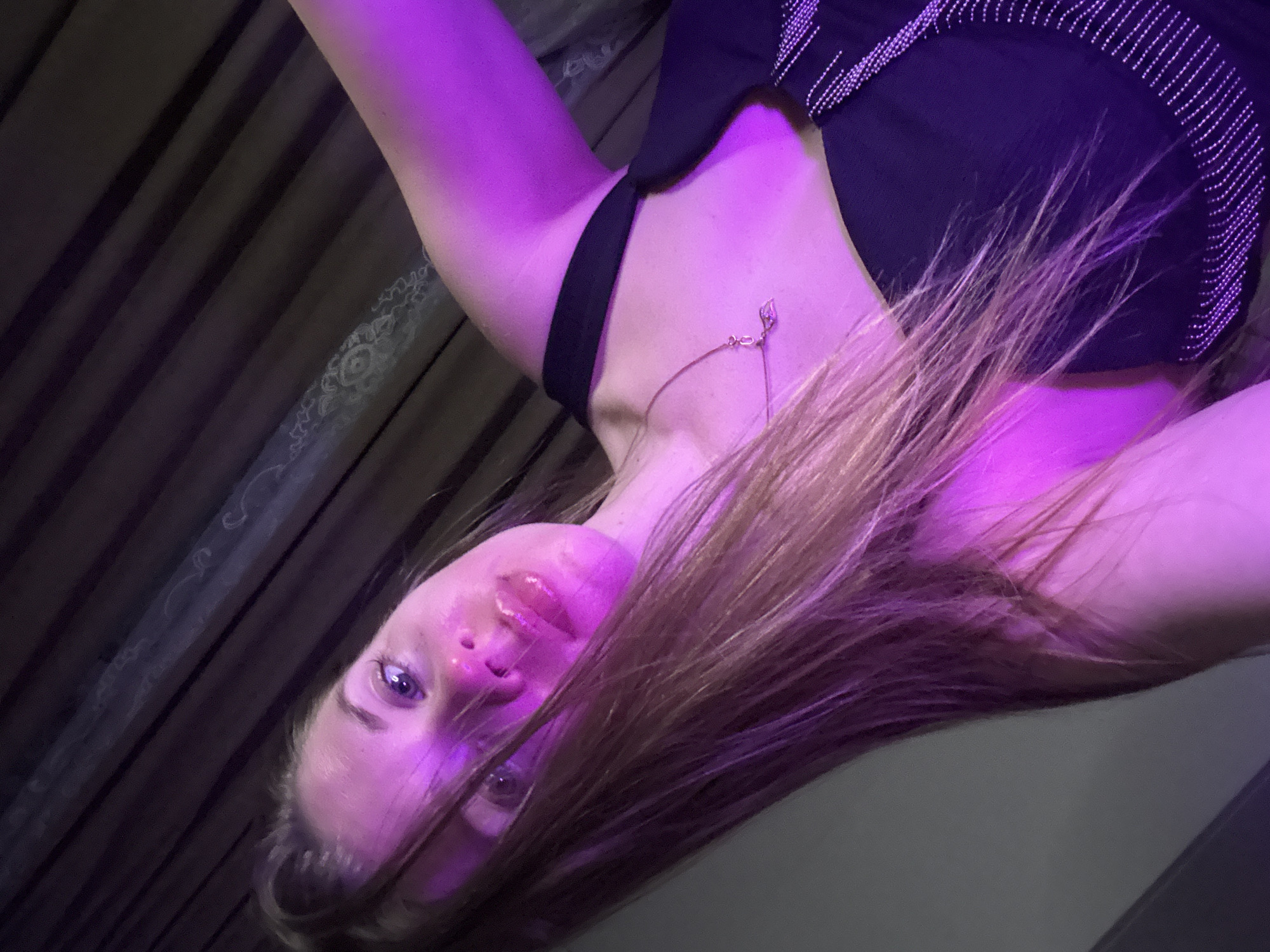A young woman is depicted in a close-up, angled portrait, illuminated primarily by strong purple and pink lighting. She is positioned from the chest up, with her head tilted downwards towards the camera, and her body is oriented diagonally in the frame. Her long, light brown hair cascades around her face, partially obscuring her left side. She has expressive eyes and glossy lips, looking directly at the viewer with a neutral to slightly pouting expression.
She wears a dark (likely black) top, which features a distinctive beaded or studded pattern along its edges. A delicate silver-toned necklace with a small, possibly leaf-shaped, pendant adorns her neck.
The background consists of dark, vertically striped fabric, possibly curtains or wall coverings, with a subtle, lighter-toned ornate pattern visible in areas. A plain, darker surface occupies the lower right portion of the image. The artificial, colored lighting creates a vibrant and atmospheric mood, suggesting an indoor setting such as a club, lounge, or a room with specialized mood lighting, likely at night. The unique angle and composition imply a stylized or artistic photographic pose. No text is visible in the image.