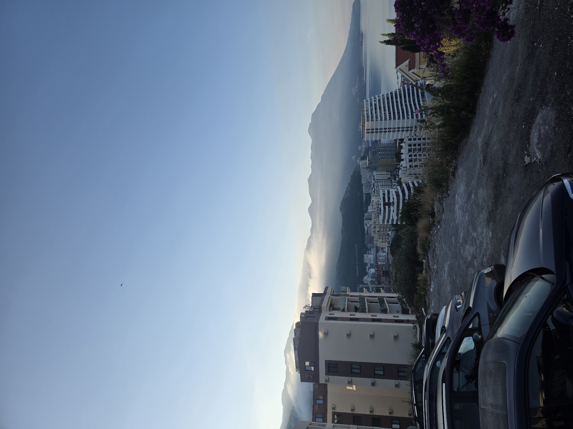 The image depicts a scenic view overlooking Bečići, Montenegro. In the foreground, a gravel parking area is occupied by several parked cars, including a dark-colored sedan in the lower portion of the frame. Beyond the parking lot, a multi-story apartment building with balconies and air conditioning units is visible. Further in the background, a dense urban landscape of various buildings, including modern high-rises, extends towards the sea. Mountains shrouded in mist form a dramatic backdrop to the city, with the calm sea visible on the right side of the frame. The sky is clear and blue, with a faint trace of a contrail or a bird in the upper left quadrant. The overall scene suggests an early morning or late afternoon ambiance due to the soft lighting.