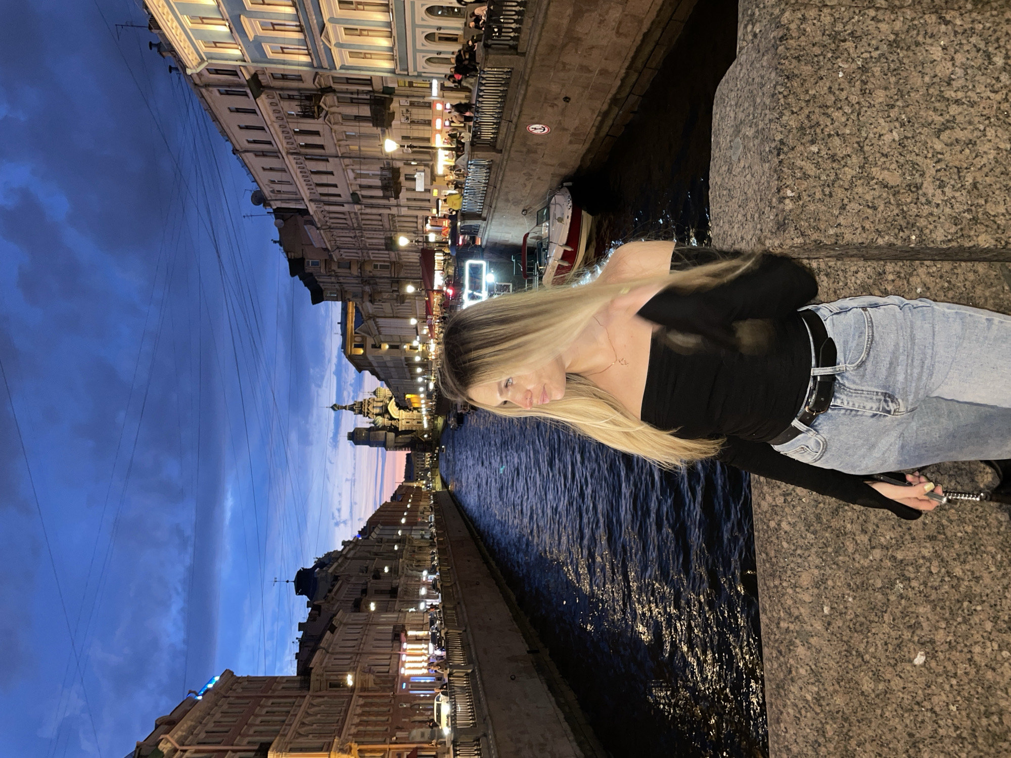 A young woman with long blonde hair, wearing a black off-the-shoulder top and light blue jeans, stands on a granite bridge overlooking a canal in Saint Petersburg, Russia. The time of day appears to be dusk, with a twilight sky overhead, transitioning from a deep blue to shades of pink and orange near the horizon. The canal is flanked by multi-story buildings with illuminated windows and decorative architecture. A red and white boat is docked along the canal. Streetlights cast a warm glow on the scene, and power lines are visible crisscrossing the sky. In the background, a distinct onion-domed church structure is visible. The overall atmosphere is that of a picturesque evening in the city.