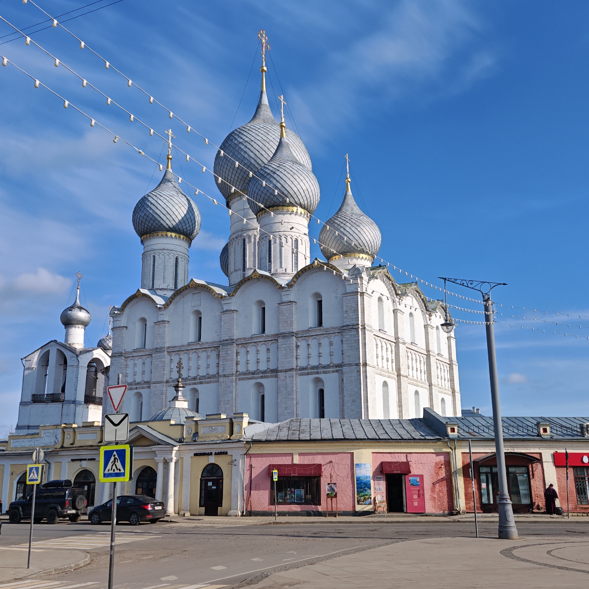 The image depicts a large, white cathedral with silver domes in Rostov, Russia. The cathedral is multi-tiered and ornately decorated. In front of the cathedral is a street with several buildings on the opposite side, including shops. A person wearing a dark robe and carrying a bag can be seen standing outside of one of the shops. There are also cars and street signs visible in the foreground. Overhead street lighting is in the upper section of the photograph. The sky is blue with sparse clouds.