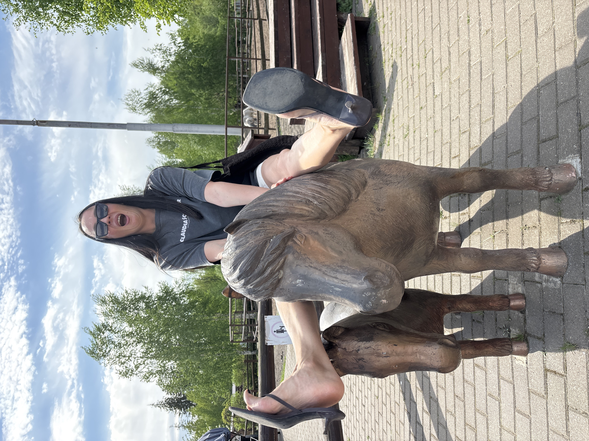 A person is straddling a bronze horse sculpture outdoors. The individual is wearing a grey t-shirt, dark sunglasses, and black high-heeled sandals. The right shoe is off, revealing the sole. The surrounding environment includes brick paving, trees, a wooden fence, and a blue sky with clouds. Based on the presence of a recognizable sculpture, this scene is in Izhevsk, Russia.