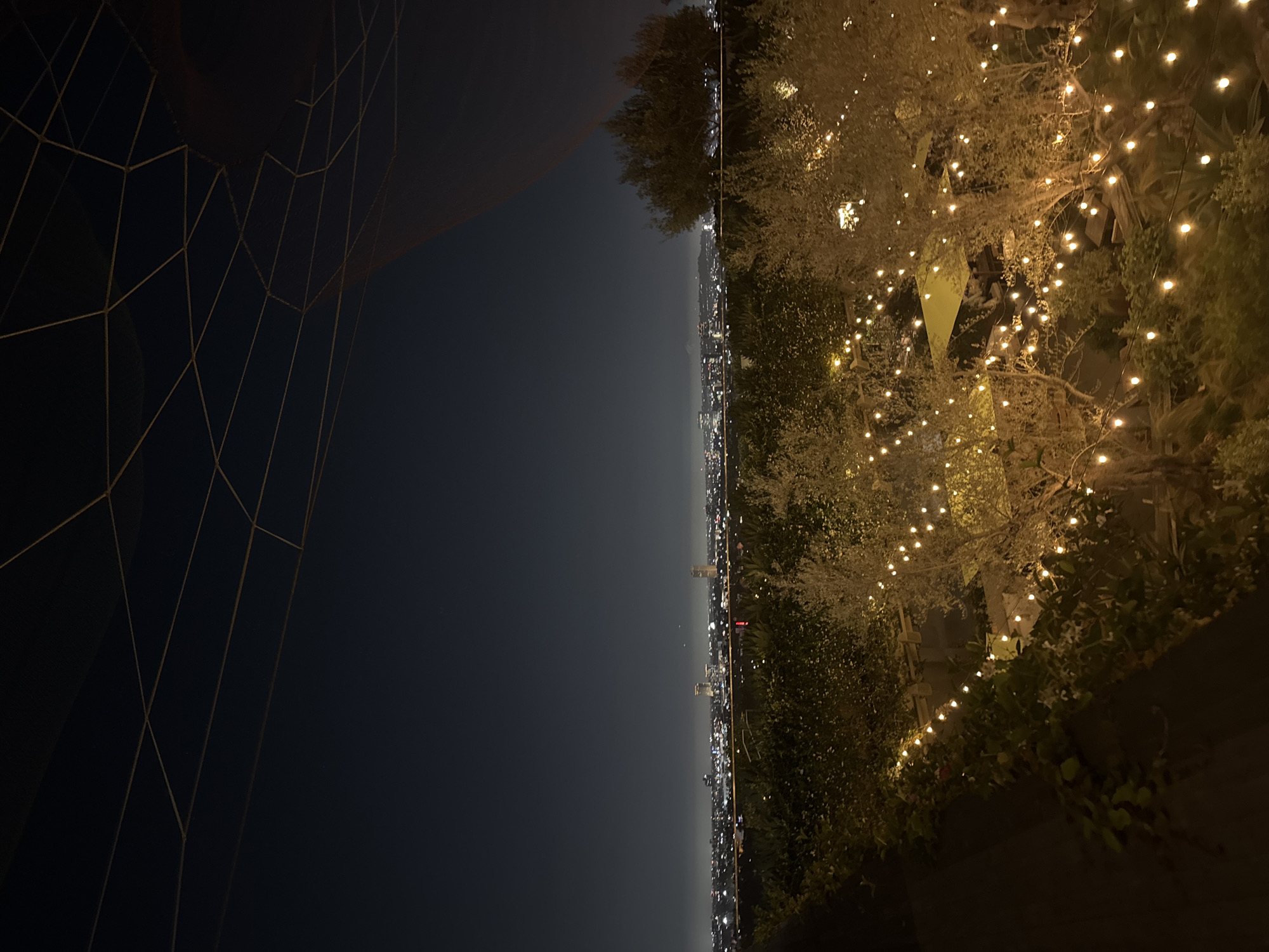 The image captures an elevated night view over West Hollywood, United States of America. On the left side, a dark, abstract architectural feature displays a prominent geometric grid pattern with golden or light-colored lines.
To the right, an outdoor space is illuminated by numerous warm, glowing string lights draped across trees and bushes, creating a festive and inviting atmosphere. A light-colored, possibly triangular or tent-like structure is faintly visible within the illuminated foliage.
In the background, a sprawling city skyline extends along the horizon under a dark sky, dotted with countless twinkling lights from distant buildings. The sky is clear, allowing for a panoramic view of the urban landscape. There are no people or discernible activities visible. The scene evokes a tranquil and sophisticated ambiance. No text is visible in the image.