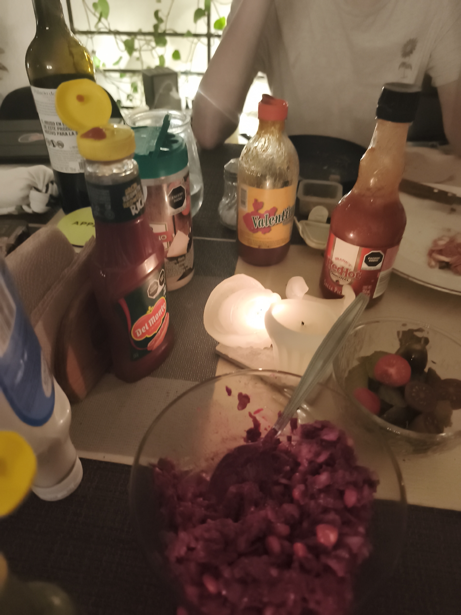 A table setting in Mexico City, Mexico, shows several food items and condiments arranged for a meal. A bottle of wine with a yellow lid is positioned near a container of spices, beside a brown napkin. Next to it is a bottle of Del Monte tomato sauce, topped with a yellow lid.
A burning candle sits near two bottles of hot sauce, one branded "Valentina" and the other "RedHot." In the foreground, two glass bowls contain food, one with a purple mixture and the other with tomatoes and green vegetables. A spoon rests in the bowl with the purple mixture.
A person is partly visible in the background, wearing a white shirt. Other elements include plates, a fork, and other unidentified objects. The setting indicates a casual dining environment.