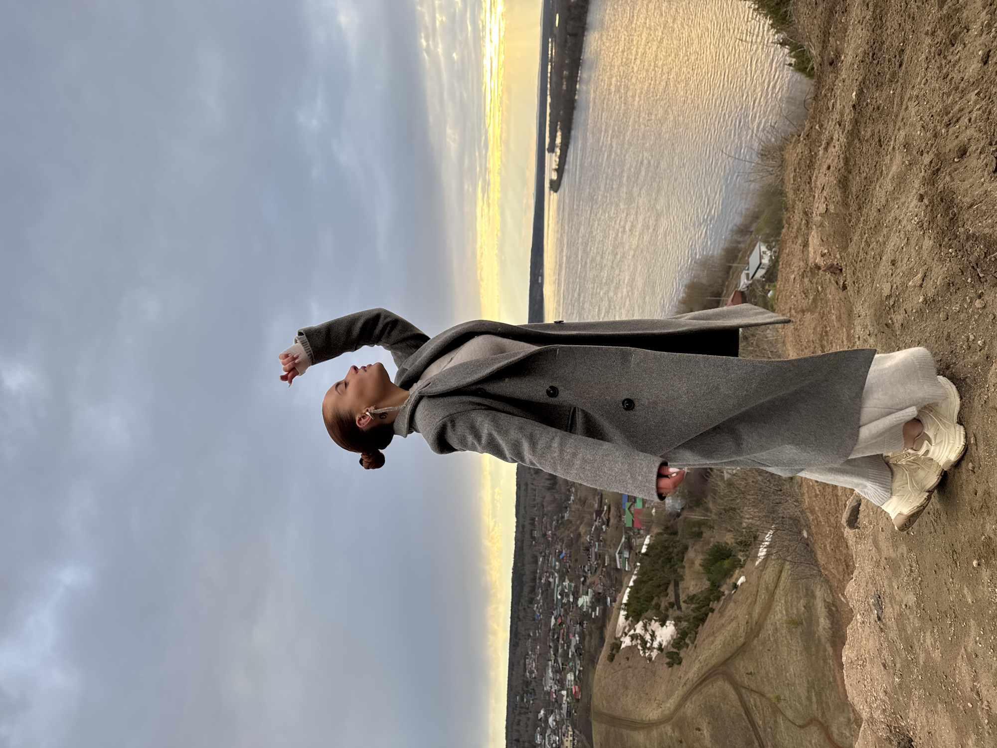 A medium-shot portrait captures a woman standing on a rugged, earthy cliff overlooking a vast river and a distant settlement, likely in Olkhovka, Russia. The woman, shown in profile, has her hair styled in a bun and wears prominent earrings. She is dressed in a long grey double-breasted coat, light-colored pants, and light-toned sneakers. Her arm is raised in a gesture that suggests contemplation or appreciation of the expansive view, as she looks upwards and slightly to the side.
The scene is set during late afternoon or early evening, possibly sunset, with a golden glow visible on the horizon and reflecting across the river's surface. The sky above is predominantly overcast with grey clouds, creating a striking contrast with the brighter water. Below the cliff, a village with numerous houses and trees is visible, with patches of snow indicating either early spring or late autumn. A winding dirt path can be seen on the hillsides leading down towards the river. The natural, uncultivated terrain emphasizes the remote and elevated location. There is no visible text in the image.
