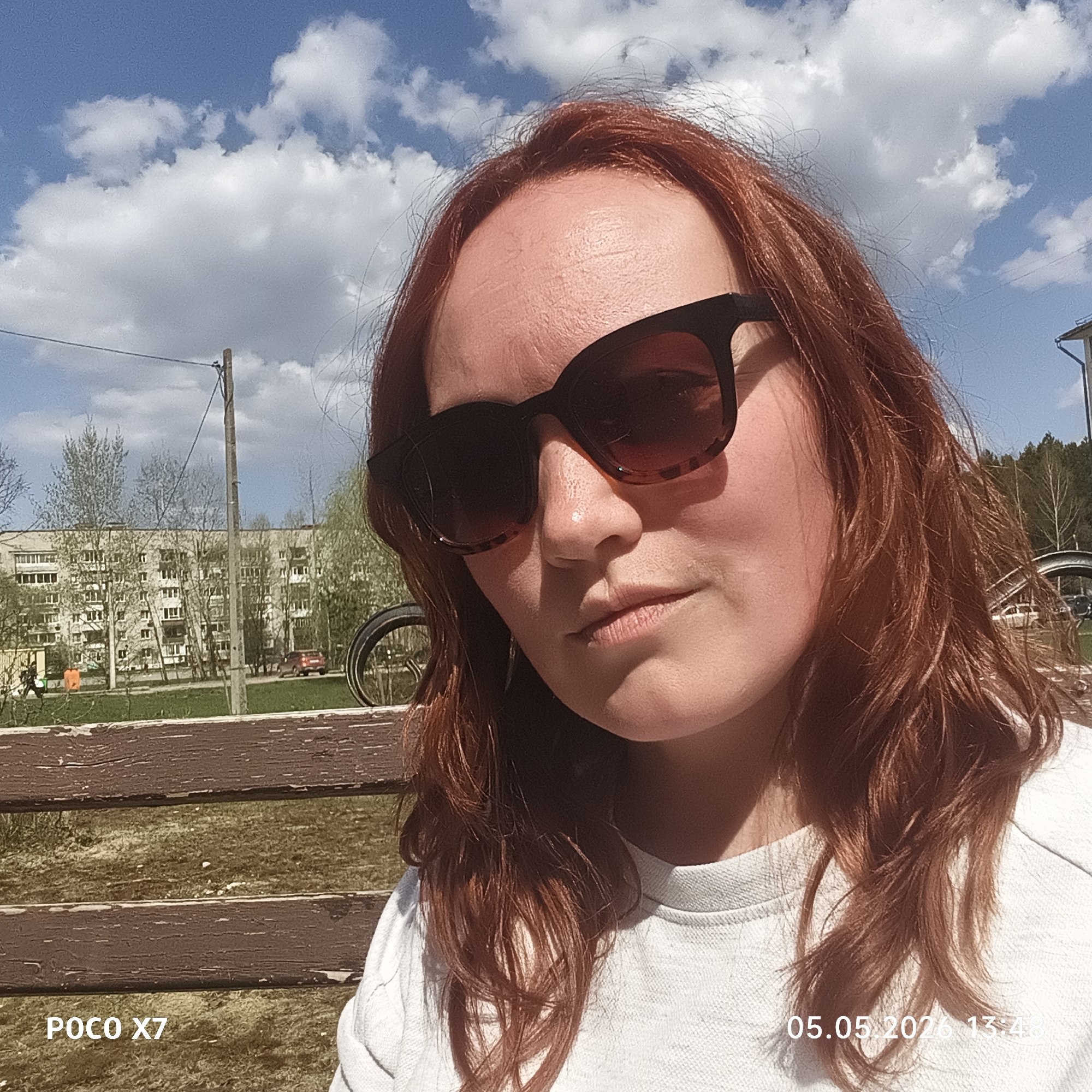 The image presents a close-up of a woman with red hair and sunglasses. She is wearing a white shirt and is outdoors on a sunny day with some clouds in the sky. Behind her is a wooden bench, a grassy area, a building, trees, and a utility pole. In the lower left corner, the text "POCO X7" is visible. In the lower right corner, the text "05.05.2026 13:48" is visible. Based on the available information, it is impossible to confirm whether the location is specifically in the city of Chaikovsky, Russia.