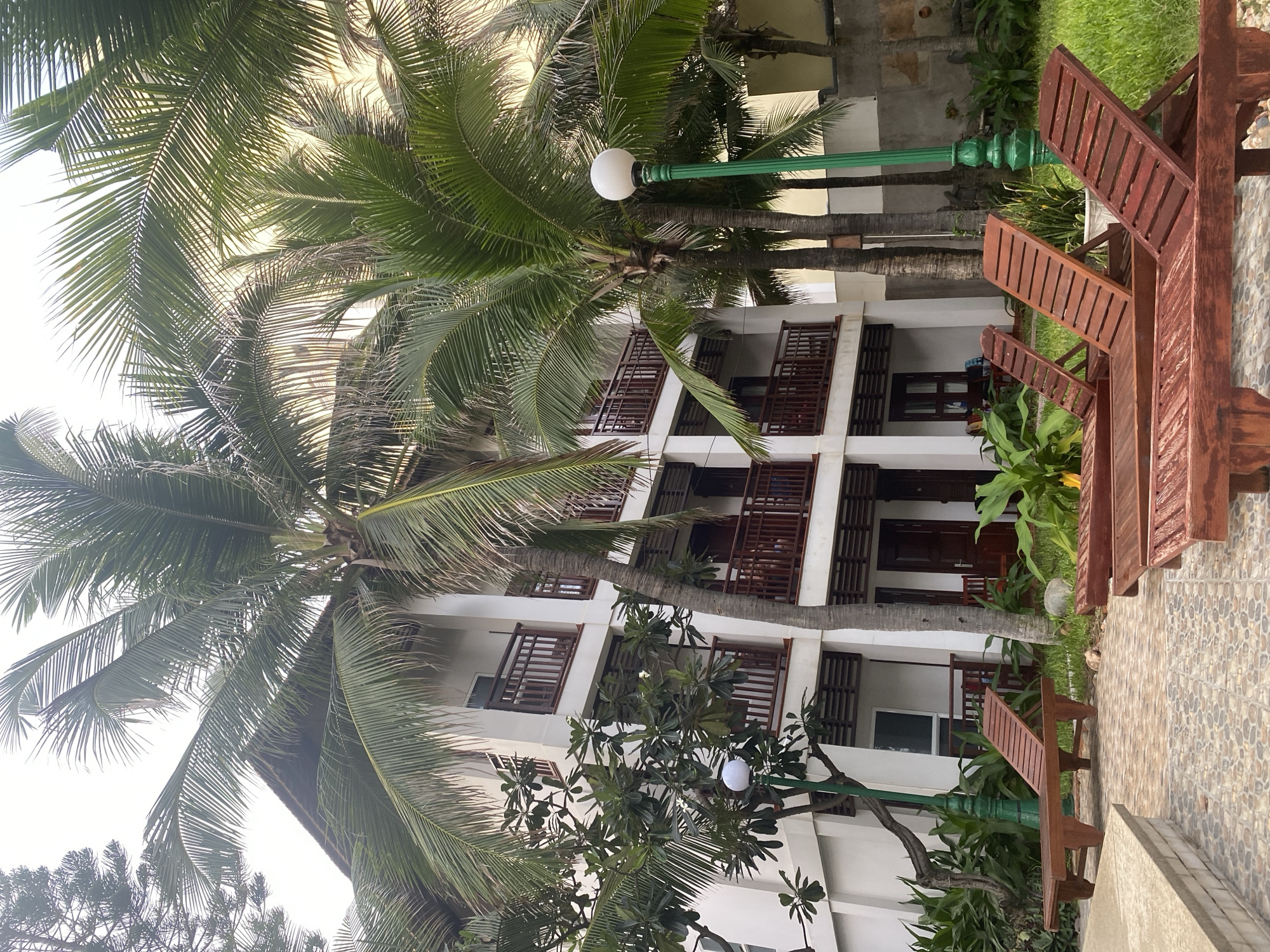 A resort building with white walls and balconies with dark brown railings is pictured. Several palm trees with green fronds are in front of the building. In the foreground, there are three wooden lounge chairs with adjustable backs. A tiled walkway is in front of the lounge chairs, and a grassy area is behind them. A green lamppost with a white globe is located to the right of the building. The image appears to be taken during the daytime.