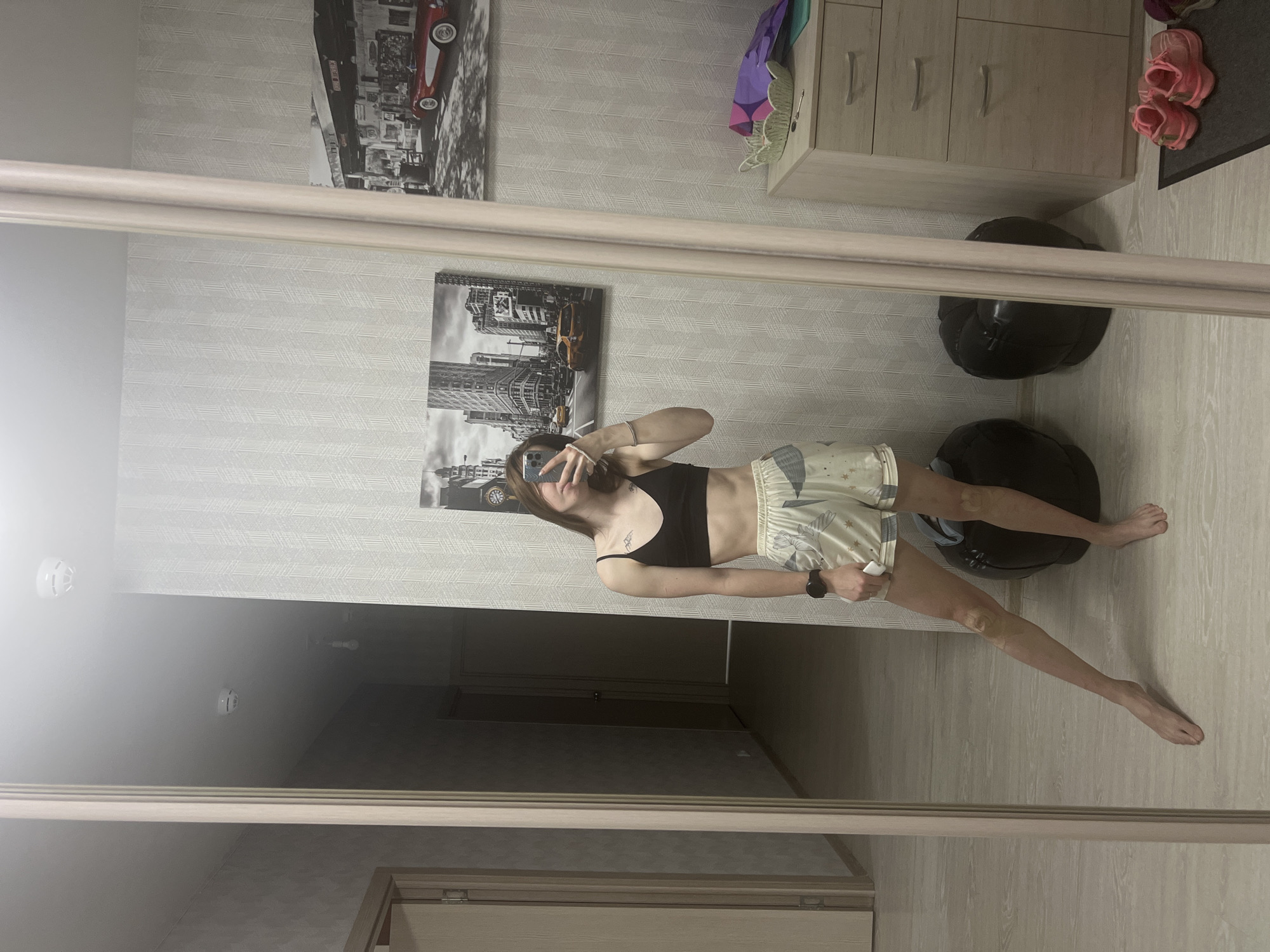 A slender, fair-skinned female with shoulder-length brown hair stands barefoot in an interior setting. She wears a black sports bra and patterned shorts. She holds a mobile phone in her left hand, obscuring part of her face. A tattoo is visible on her upper left torso.
Behind her hangs two black and white pictures on the wall, the top one featuring cars and buildings, the bottom one featuring a city skyline with a yellow taxi cab. To the right is a light-colored dresser with closed drawers. Also to the right are pink sandals and two black weights.
The setting includes flooring that resembles light-colored wood. The photograph appears to be a reflection in a mirror or other reflective surface, as the walls behind the woman feature repeating wallpaper. The background also shows a partially obscured doorway and ceiling.
There is insufficient information within the photograph to determine the specific location in Perm, Russia.