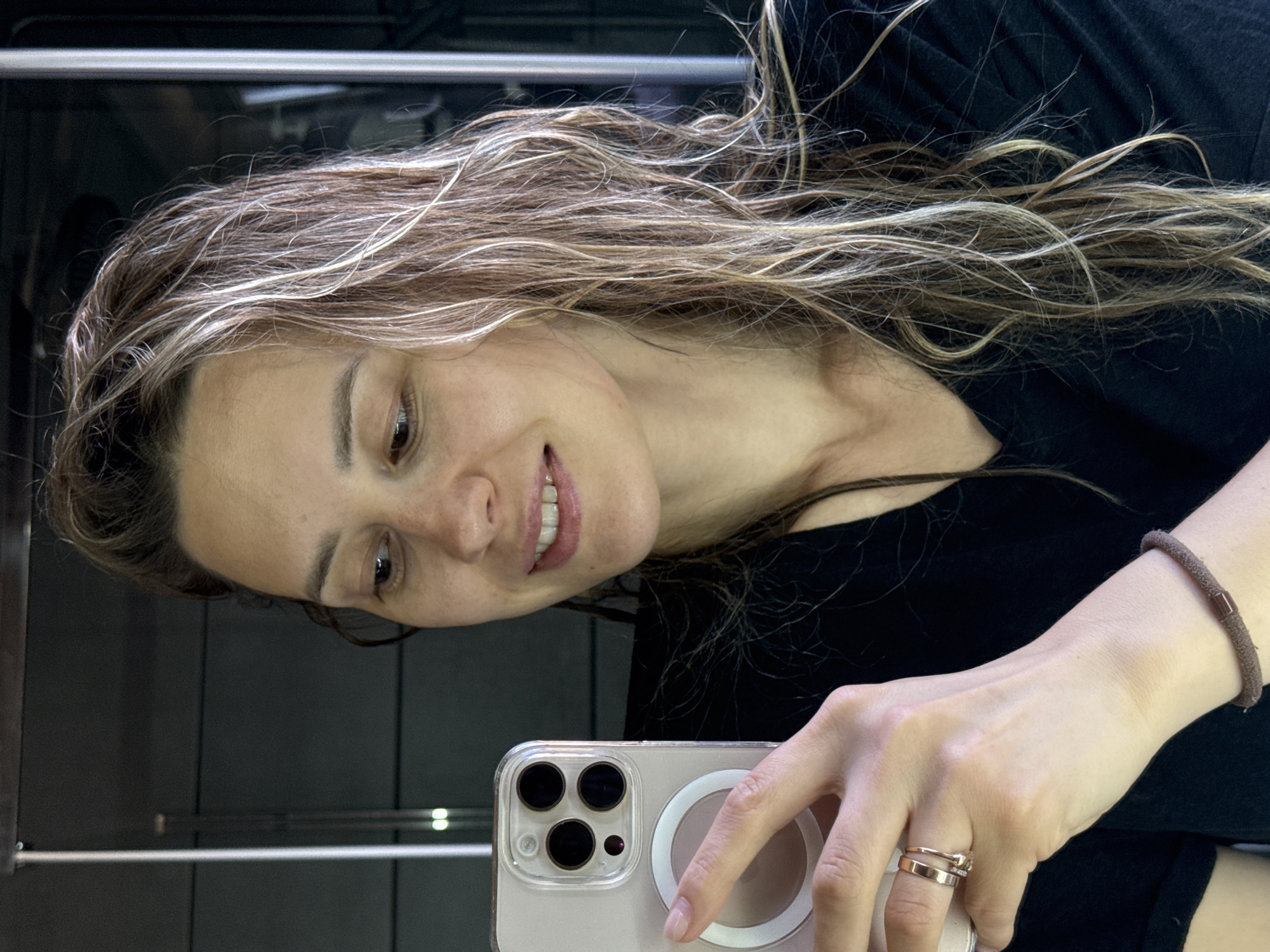 A woman with highlighted brown hair and a black shirt is holding a smartphone with a clear case and a MagSafe charging ring. She is looking at the phone with a slight smile. Her left hand is visible, adorned with two gold rings and a brown hair tie around her wrist. She is positioned in front of a mirrored surface, possibly an elevator, reflecting the surroundings. There is no information available to identify that the picture was taken in the city of Chaikovsky, Russia.