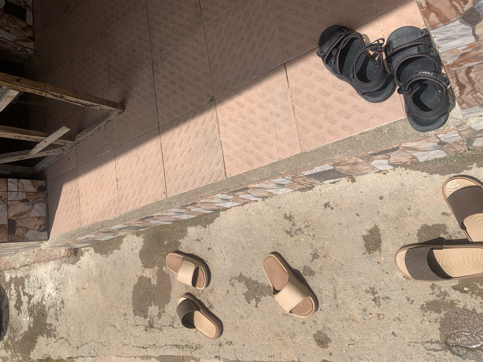 In Jauro Yinu, Nigeria, a collection of footwear is scattered on a wet concrete patio adjacent to a tiled walkway. Two pairs of beige slides are visible, one positioned near the top left and another further down. A third pair of slides, with olive green straps, lies next to the top beige pair. Towards the right side of the image, on the tiled walkway, two pairs of black sandals are placed. The scene appears to be outdoors, with sunlight casting shadows.