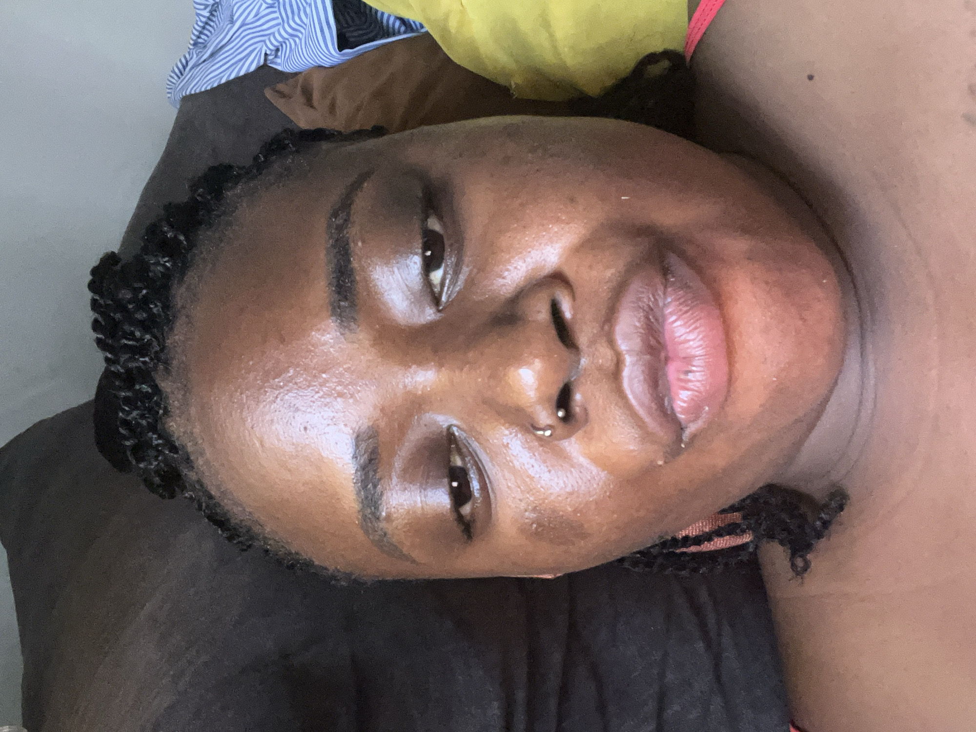 The image is a close-up selfie of a Black woman, likely taken indoors. Her head is tilted slightly back, and her eyes are partially closed. She has dark, braided hair. There's a nose ring visible. Her skin appears shiny, possibly from sweat or skincare products. She is wearing a pink garment. In the background, there is a dark surface, possibly a sofa or bed, and a glimpse of blue and white striped fabric, suggesting clothing or bedding. There is also a bright yellow object to the right, which could be a pillow. The lighting suggests it is daytime. The image does not provide any information about the city of Abuja, Nigeria, or any specific location within it.