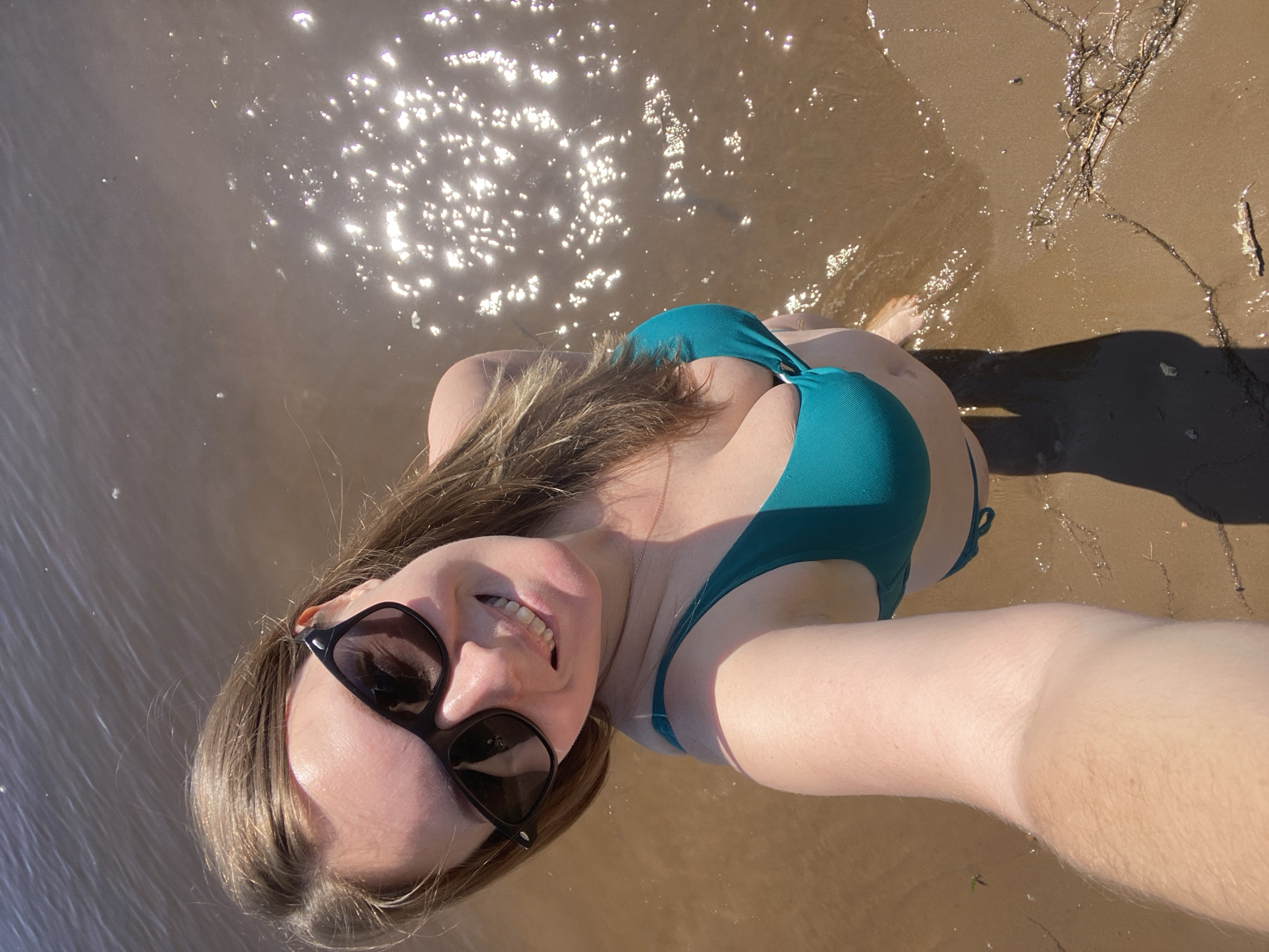 The image shows a woman in a teal bikini standing in shallow water at the edge of a sandy beach. She is wearing black sunglasses and smiling at the camera, which is positioned above her, taking a selfie. Her hair is light brown and falls around her shoulders. The water is calm and reflects the sunlight, creating a shimmering effect. The beach is made of sand and small pebbles. The overall scene suggests a sunny day spent at a body of water. It is not possible to determine the specific location within the city of Chaikovsky, Russia from the image alone.