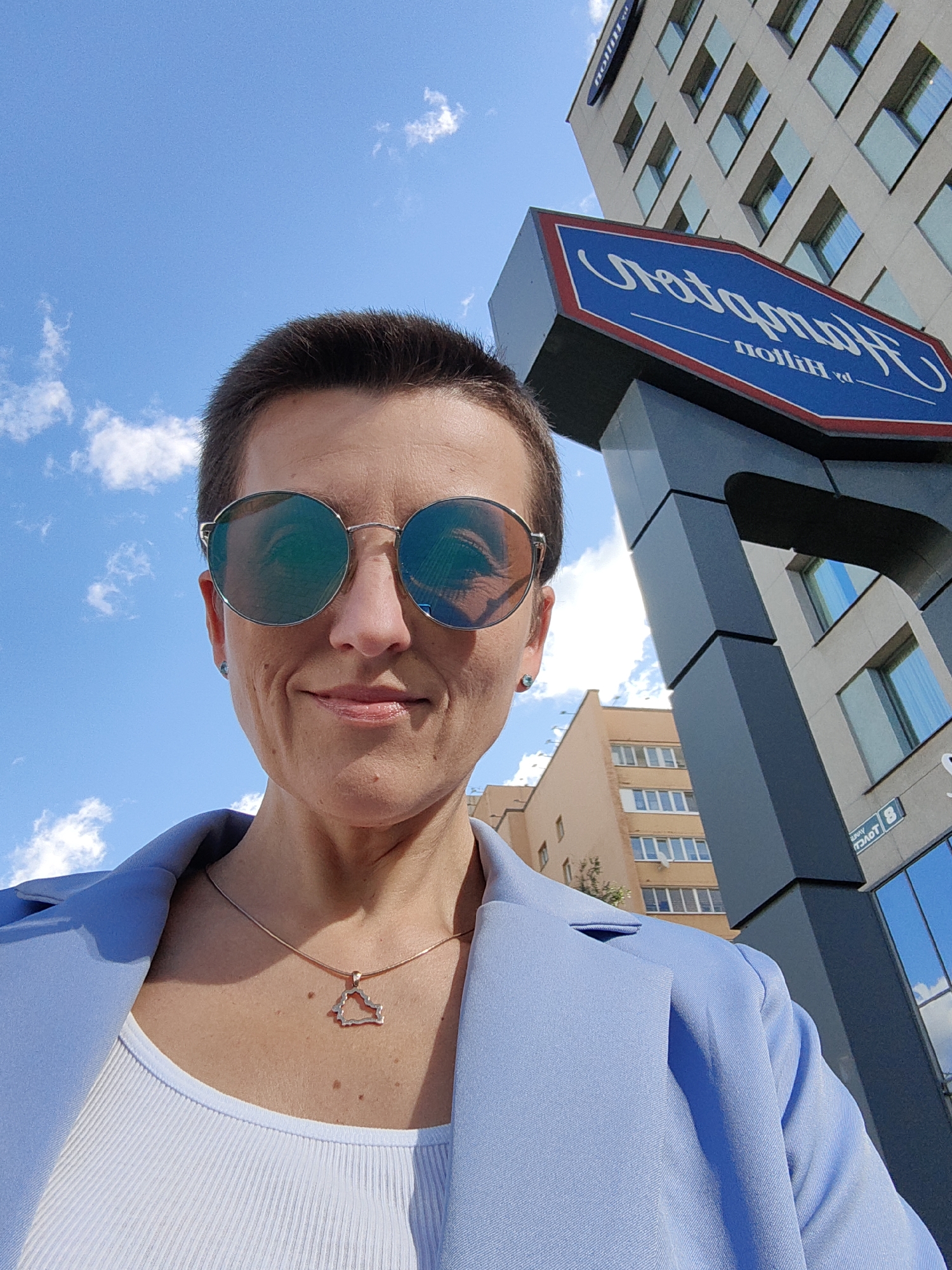 A low-angle, medium shot shows a woman with a short haircut and sunglasses looking at the camera, with a sky and a building in the background. The woman is wearing a light blue blazer over a white ribbed tank top and a rose gold chain with a pendant shaped like the outline of Belarus. The building in the background is a multi-story structure with many windows, and a prominent blue sign with white text reads "Hampton by Hilton." The sky is a bright blue with scattered white clouds. The image appears to be taken outdoors in an urban environment, likely in front of the Hampton by Hilton hotel in Minsk, Belarus.