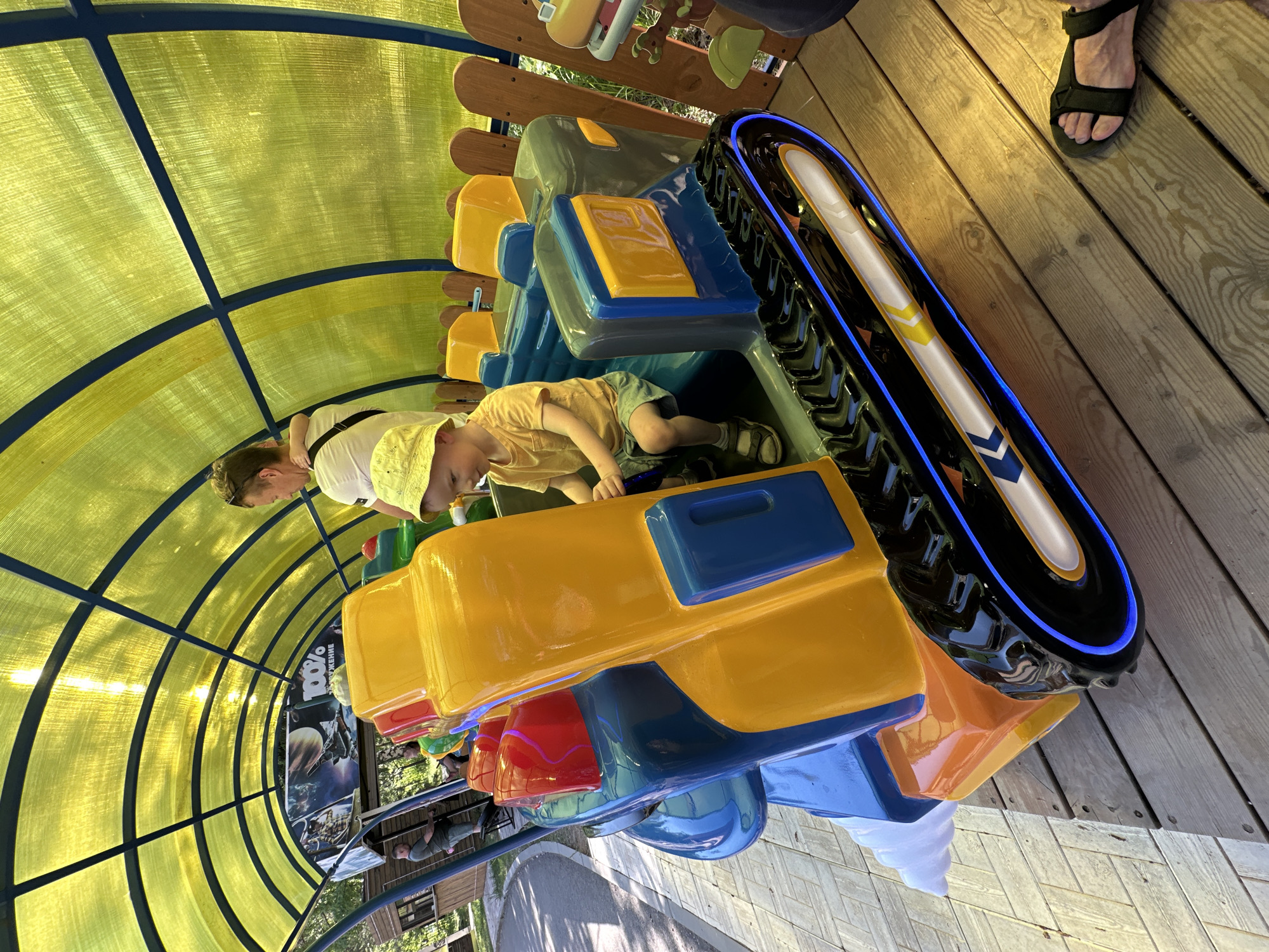A color photograph shows a small boy sitting in a colorful children's train ride at an amusement park, presumably located in the city of Chaikovsky, Russia. A woman, likely his mother, stands beside the train, watching the boy. The train is covered by a yellow, translucent tunnel, which curves overhead. The train itself has yellow, blue, red, and black colored cars. The boy is wearing a yellow shirt and green shorts, and is looking down. The woman is wearing a white shirt and a yellow hat. The ride is positioned on a wooden platform.