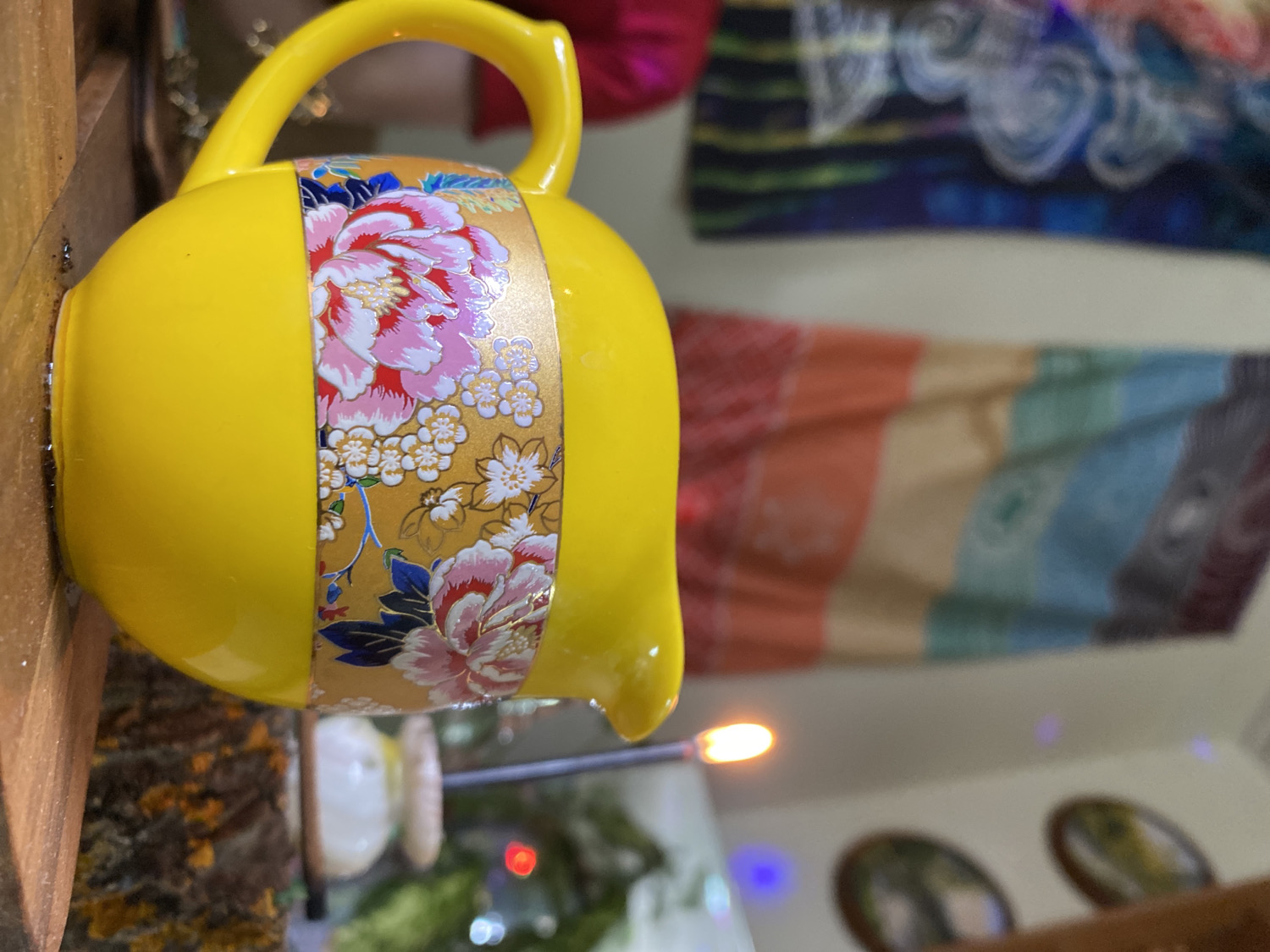 The image features a vibrant yellow ceramic pitcher or teapot, lying on its side, resting on a textured wooden surface. The ceramic piece has a prominent handle and a small spout. It is adorned with a metallic gold band wrapping around its center, intricately decorated with large, lush pink and white peony-like flowers, smaller white blossoms, and blue-green foliage. The floral pattern gives it an elegant, possibly Asian-inspired, aesthetic.
The background is softly blurred, indicating a shallow depth of field. In the upper right, a dark fabric with swirling patterns in shades of blue and white is visible. Below it, another blurred fabric, possibly a blanket or tapestry, displays horizontal stripes in warm tones of orange, beige, and light blue. In the lower center, a bright, small flame, likely from a candle, casts a warm glow. Nearby, blurred circular lights in blue and red suggest additional decorative lighting or bokeh effects. Further to the lower right, two oval-shaped objects, possibly framed pictures or small mirrors, are discernible on a wall.
The setting appears to be indoors, likely a home or a shop, characterized by decorative items and soft lighting. No people are visible, and there is no explicit activity taking place beyond the display of the objects. The general atmosphere is warm and decorative. There is no visible text in the image. The location is Chaikovsky, Russia.