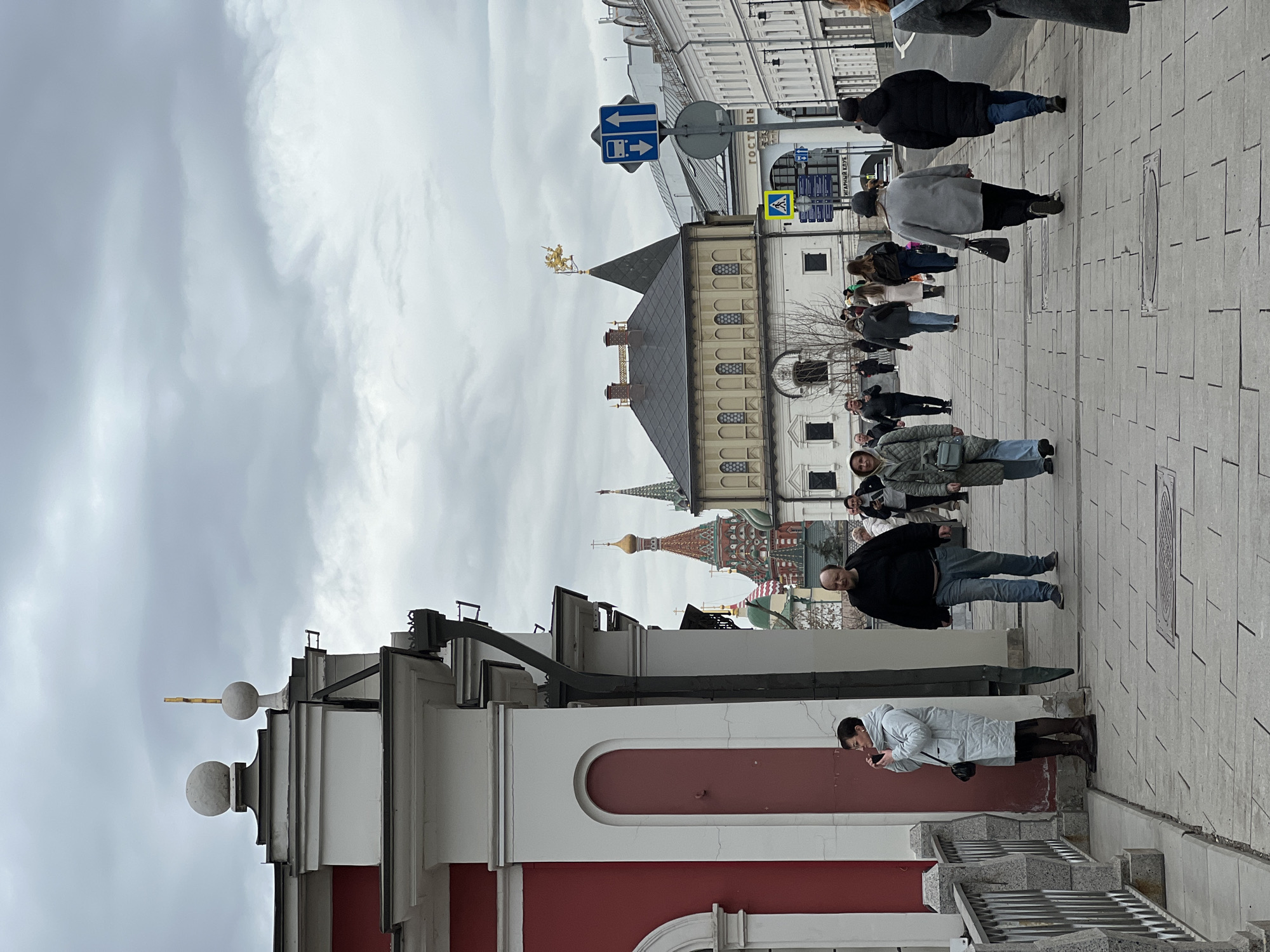 The image depicts an urban street scene on Varvarka Street in Moscow, Russia, with the Chambers of the Romanov Boyars and St. Basil's Cathedral visible in the background. The sky is heavily overcast with a uniform layer of grey clouds.
In the immediate foreground to the left, a building with a red base and white upper sections, featuring an arched niche and a dark downspout, partially frames the view. Adjacent to it is a grey stone balustrade.
The central and mid-ground comprise a paved pedestrian walkway extending into the distance, populated by approximately 15 individuals. A woman with short dark hair, wearing a light grey quilted coat and dark boots, stands near the left building, holding a small dark object to her mouth. Other pedestrians are walking in various directions; a man in a black sweater and blue jeans walks towards the viewer, while most others are receding into the background.
In the mid-ground, to the right of the walkway, stands the historic Chambers of the Romanov Boyars, a two-story building with a light-colored facade, dark grey roof, and a spire topped with a golden griffin-like sculpture. Further beyond and slightly to the left, the distinctive colorful onion domes and spires of St. Basil's Cathedral are partially visible. To the right of the Chambers, a multi-story white building with numerous windows lines the street.
Street furniture includes a tall grey pole with a blue rectangular sign indicating a bus stop and straight-ahead arrows, and a yellow diamond-shaped pedestrian crossing sign. Russian text for "ГОСТИНИЦА" (Hotel) and "КИТАЙ-ГОРОД" (Kitay-Gorod) is visible on a sign further down the street. Several manhole covers are embedded in the paved surface.