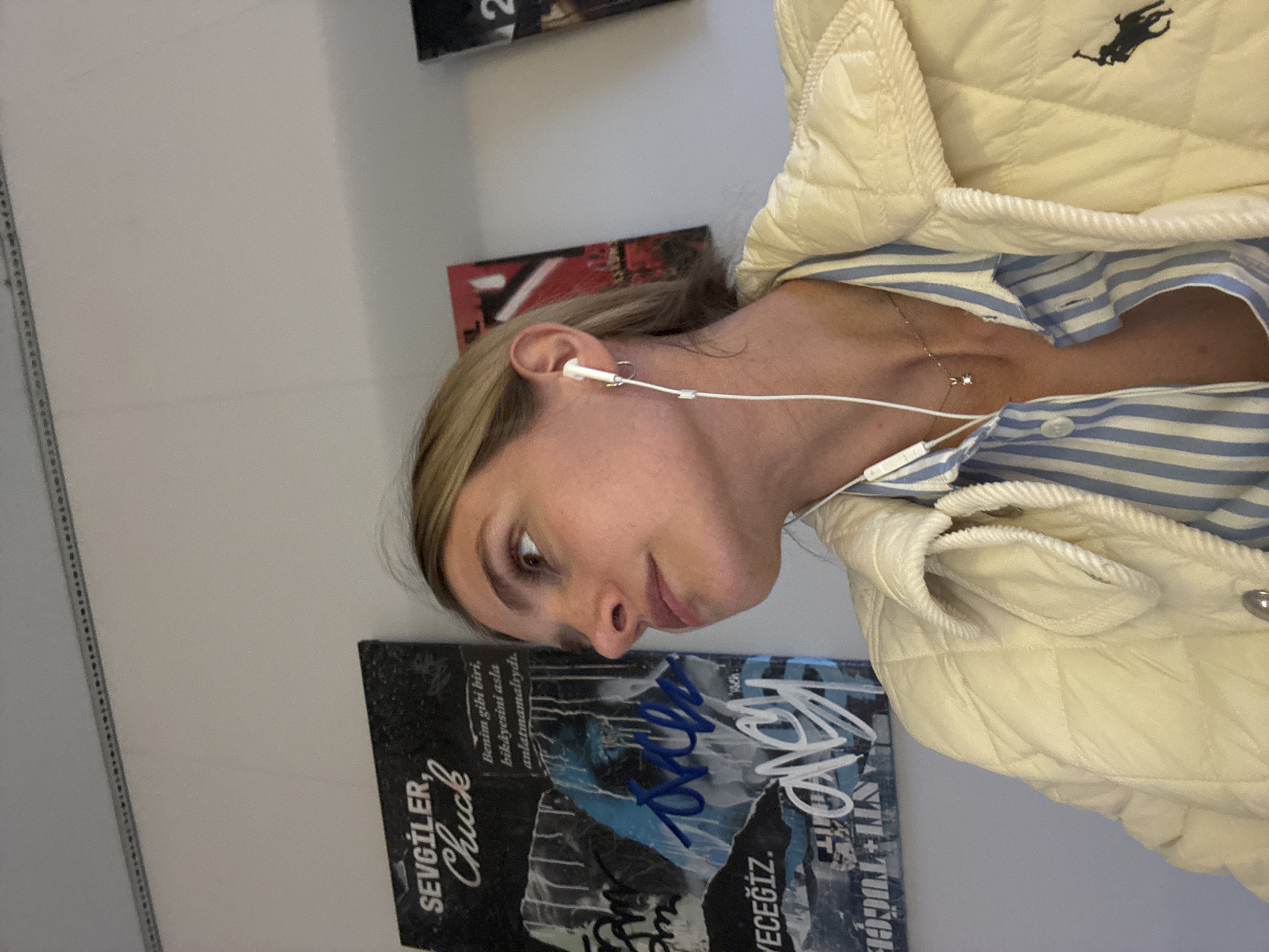 A candid, angled close-up shot features a person, likely a young adult, with light brown hair pulled back, fair skin, and brown eyes. They are wearing a cream-colored quilted jacket with a dark Ralph Lauren polo player logo on the upper right side, over a blue and white striped button-down shirt. A delicate silver-toned necklace with a small sparkling pendant rests on their chest. White wired earbuds are visible in both ears, suggesting they are listening to audio. The person's gaze is directed downwards and to the left, with a contemplative or slightly pensive expression.
The setting appears to be indoors, against a plain, light-colored wall. Several artworks or posters are affixed to the wall. The most prominent one, positioned behind the person's head, is a dark-toned poster with a collaged or graffiti-like design in blues and blacks, featuring white and blue handwritten signatures.
Visible text on this poster includes "SEVGİLER, Chuck" (Greetings, Chuck), and a Turkish sentence "Benim gibi biri, hikâyesini asla anlatmamalıydı." (Someone like me should never have told his/her story.). Partially visible text includes "VECEĞİZ." and "NTI-TUĞÇE". Another poster higher up on the wall shows the number "2". The location context provided is Tbilisi, Georgia.