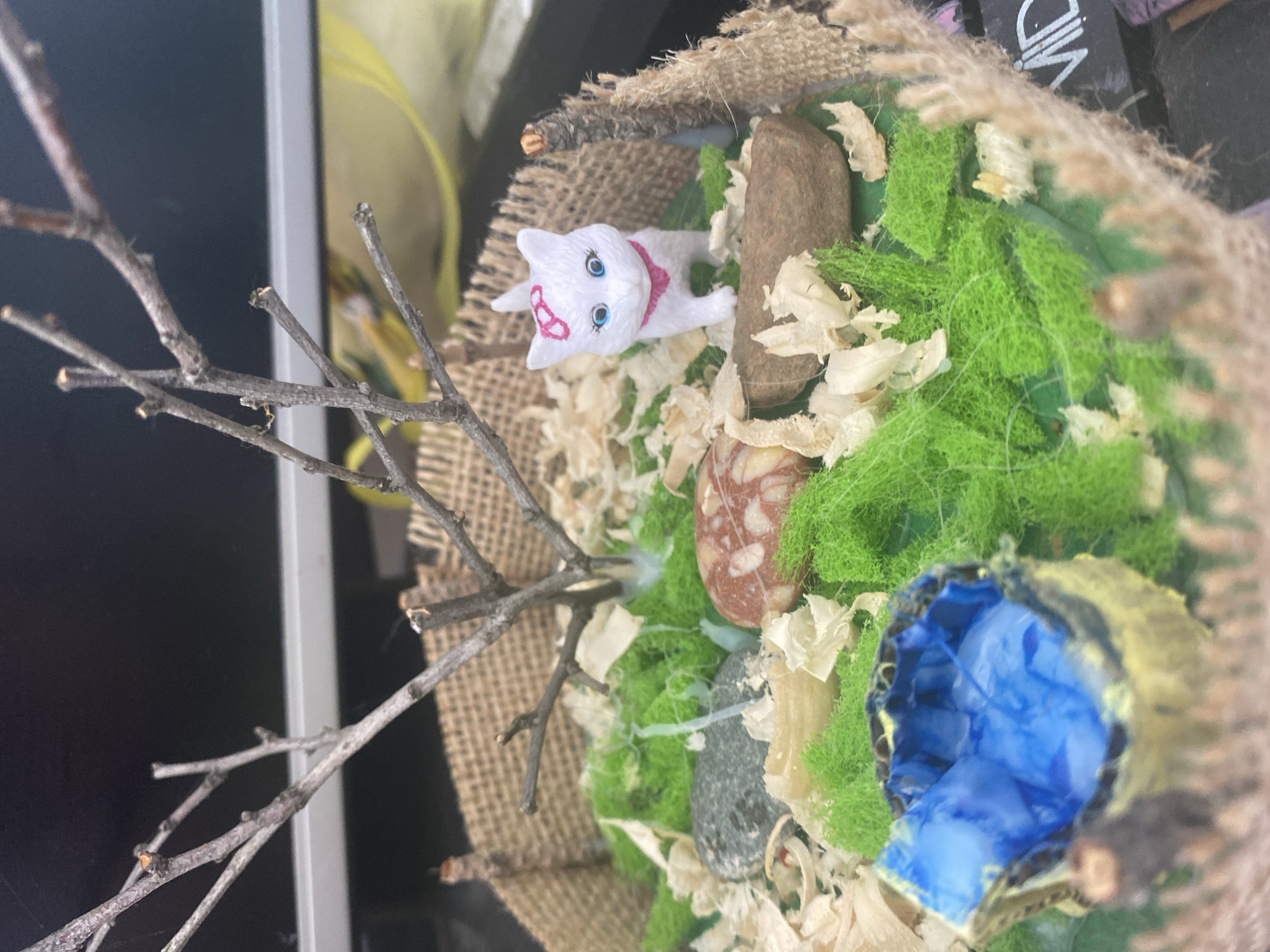 This image shows a miniature diorama featuring a white plastic cat figurine with a pink bow and collar, standing amongst green moss, wood shavings, and decorative stones. A small, blue, cup-like object is also present, appearing to represent water. A bare tree branch extends upwards from the center of the diorama. The diorama is enclosed by a rustic border made of burlap and small twigs. The background is dark and out of focus, suggesting an indoor setting. There is no indication of the city of Blagoevgrad, Bulgaria, in the image.