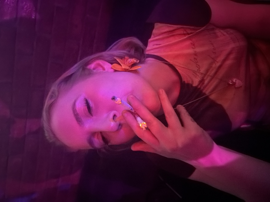 A young woman reclines under pink and purple lighting. She holds a lit cigarette in her left hand near her face, while a flower adorns her hair near her ear. Her eyes are closed, and a glittery detail is visible on her cheek. She wears a dark-collared top with a lighter-colored patterned overlay, and a necklace is visible. The background appears to be textured, possibly brick or paneling, also bathed in pink light.