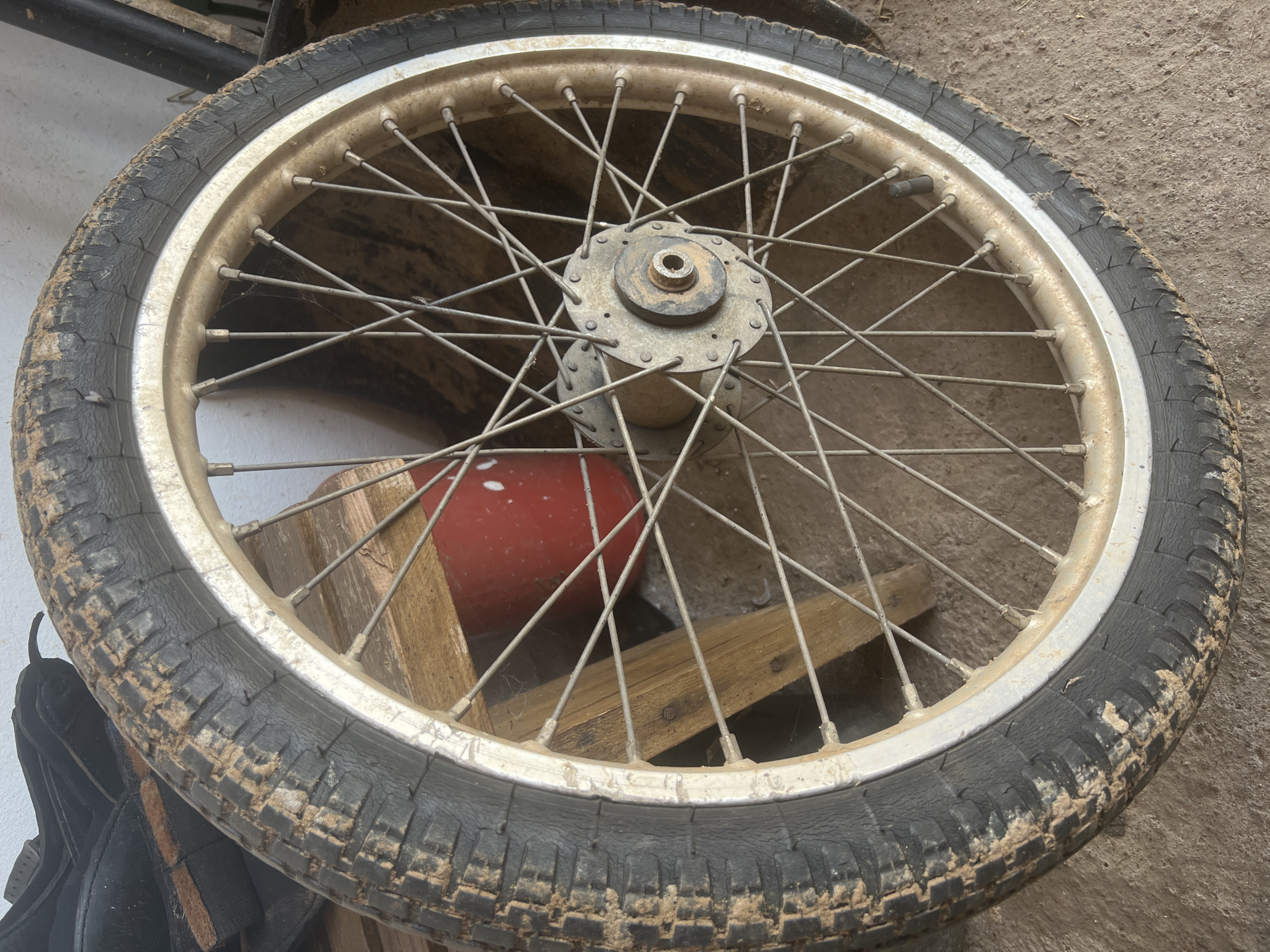 An unmounted spoked wheel, likely from a motorcycle or bicycle, lies on a dirt floor indoors. The tire is heavily soiled with dry mud. The rim is a light color, possibly aluminum or painted metal, with some discoloration and dirt. The spokes are thin and metallic, with a central hub that is also dirty and shows signs of wear. Behind the wheel, wooden planks are visible, along with a red object and a portion of what appears to be a black shoe or boot. The background is indistinct and appears to be an interior space with a wall. The scene is consistent with a storage area or workshop in Sorochinsk, Russia.