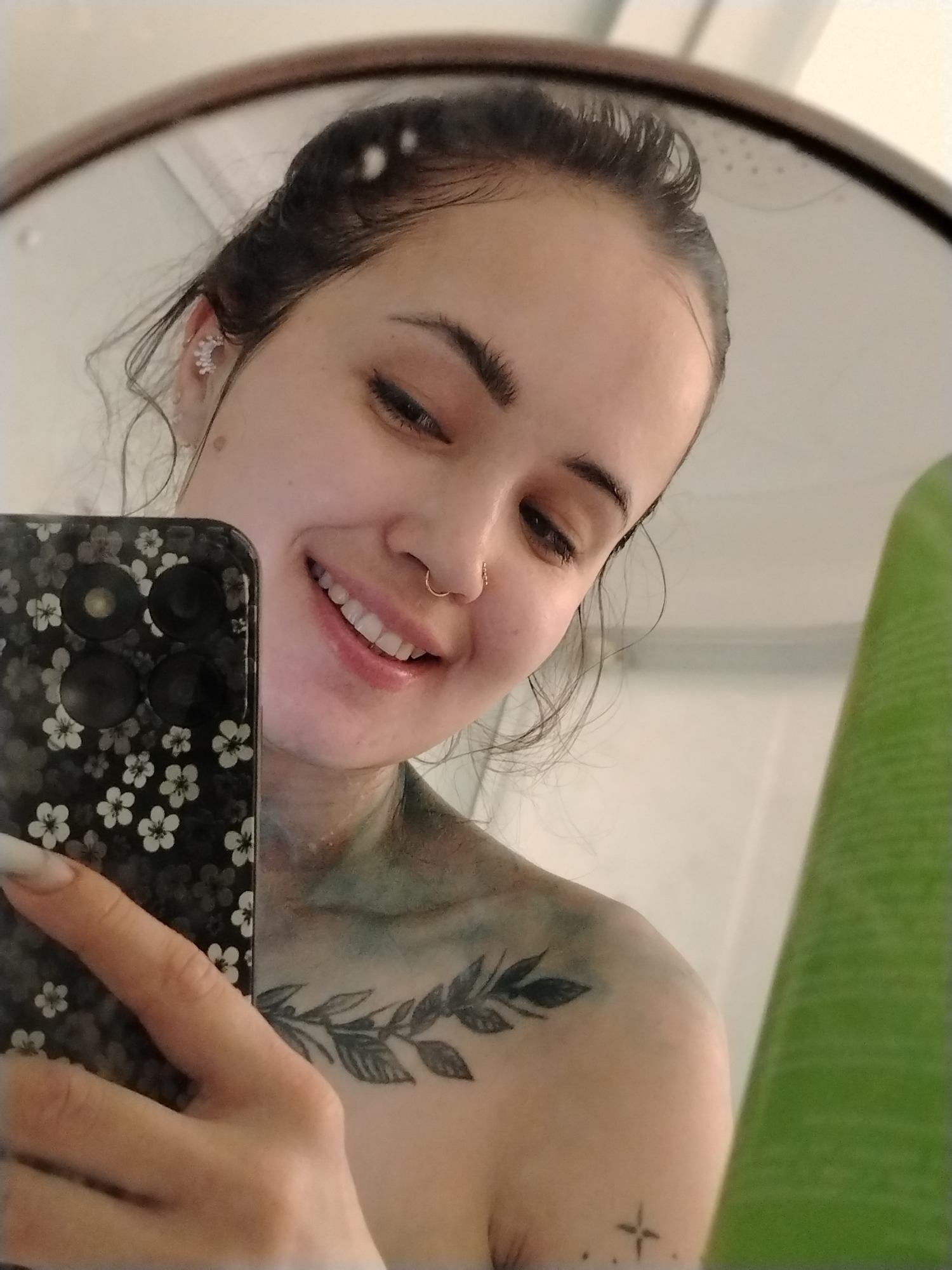 The image depicts a young woman with dark hair and fair skin taking a selfie in a mirror. She is smiling and looking down slightly. She has a nose ring and an earring. A cell phone with a floral pattern is in her hand, partially obscuring her face and upper body. A plant is visible on the right side of the image. The woman has a tattoo of leaves across her collarbone, a tattoo of a cross shape on her shoulder and some green pigment. The background appears to be a bathroom. There is no mention of the city Noviy, Russia.
