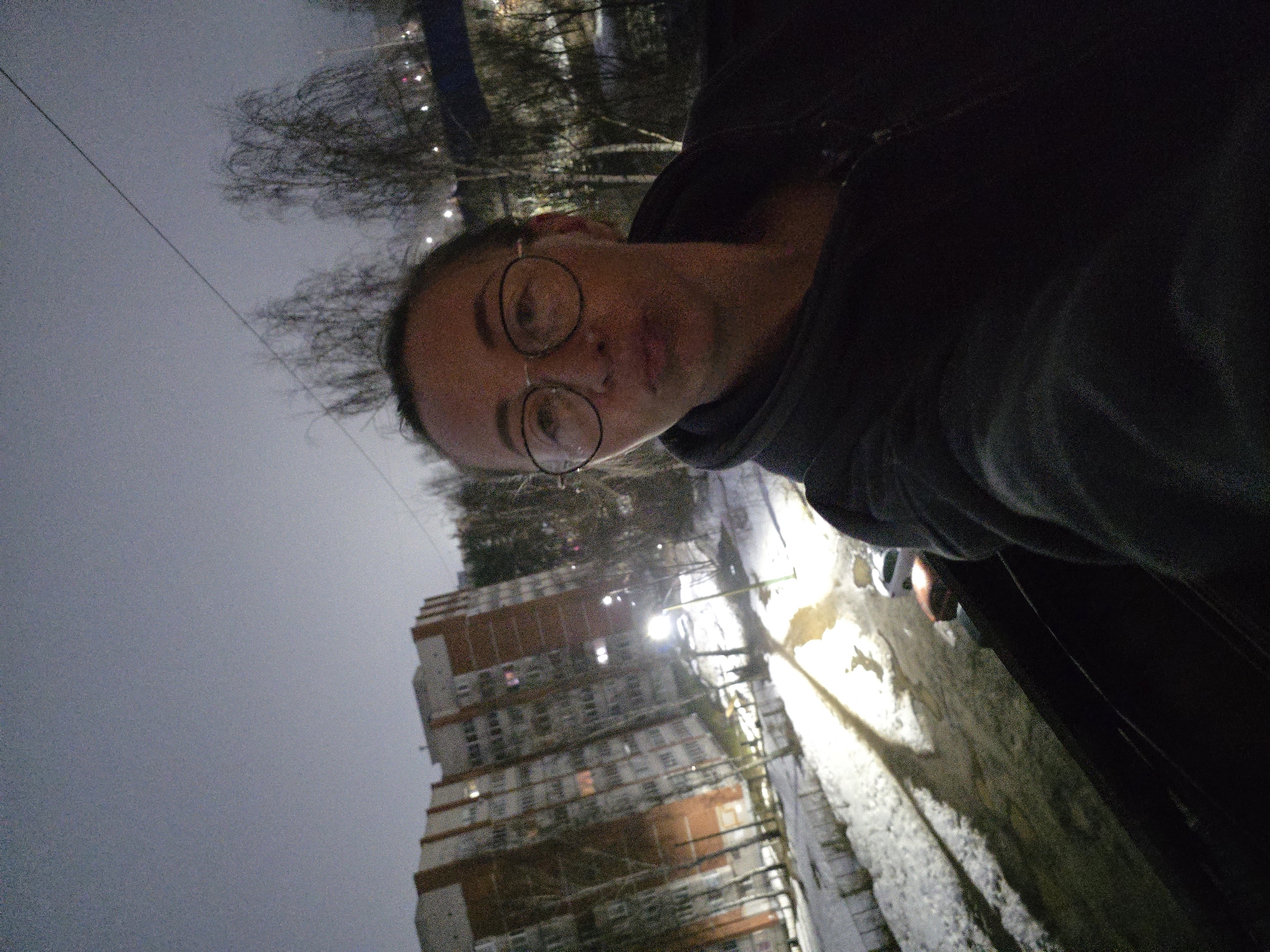 A woman with glasses is pictured outdoors at night in what appears to be Chaikovsky, Russia. She is wearing a dark hooded sweatshirt and holding the camera. In the background is a tall, brick-and-concrete apartment building with lit windows. There is some snow on the ground, and a street lamp illuminates a puddle. Bare trees are visible in the background near the top of the frame. A wire crosses the upper portion of the image.