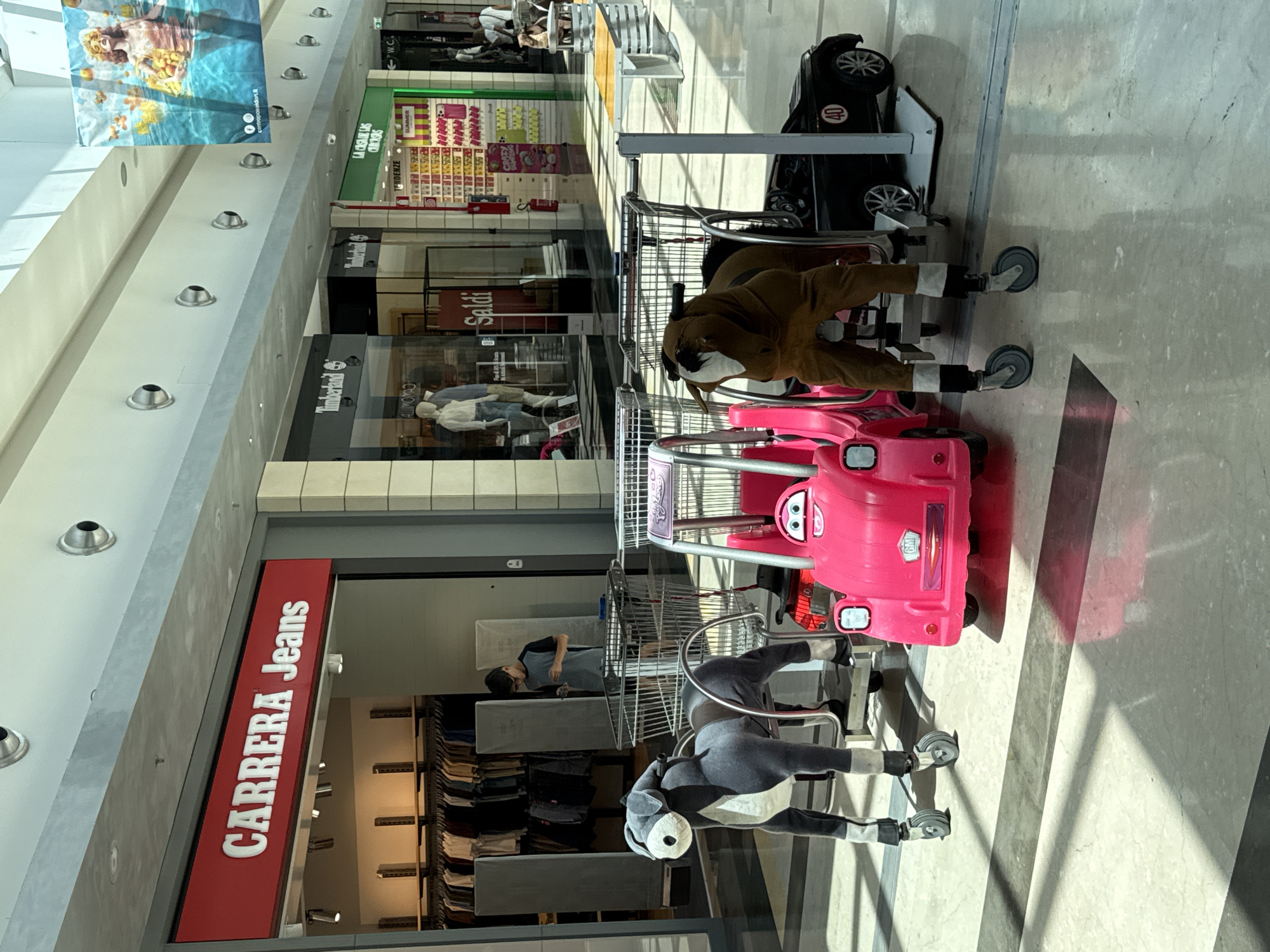 The image depicts the interior of a shopping mall, likely in Carini, Italy, with a focus on the entrance of a "CARRERA Jeans" store. In the foreground, several amusement rides for children are present. These include a grey stuffed donkey on wheels, a pink ride-on toy car resembling a stylized vehicle with cartoon eyes, and a brown stuffed horse on wheels, partially obscured by the pink car and a black ride-on toy car with the number 40 on its side. The rides are situated in front of shopping carts. A young boy in a grey t-shirt and dark shorts is standing behind the donkey ride, looking at his phone. The mall floor is tiled with a geometric pattern of light and dark squares. In the background, the "CARRERA Jeans" store is visible, along with other storefronts labeled "Timberland" and "LA CASA DEGLI OROLOGI." Sunlight streams in from above, creating shadows on the floor. A large banner featuring a woman in a swimsuit is hanging from the ceiling in the upper right portion of the image.