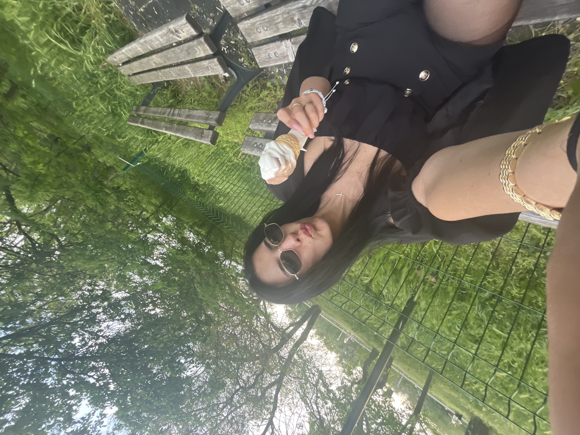 A person is sitting on a wooden bench in a park in Plovdiv, Bulgaria, holding an ice cream cone. The individual is wearing a black dress with gold buttons, black tights, and sunglasses. They are also wearing jewelry, including a gold bracelet and rings. The bench is situated on a grassy area with a green fence in the background. Trees and sky are visible in the upper part of the image.