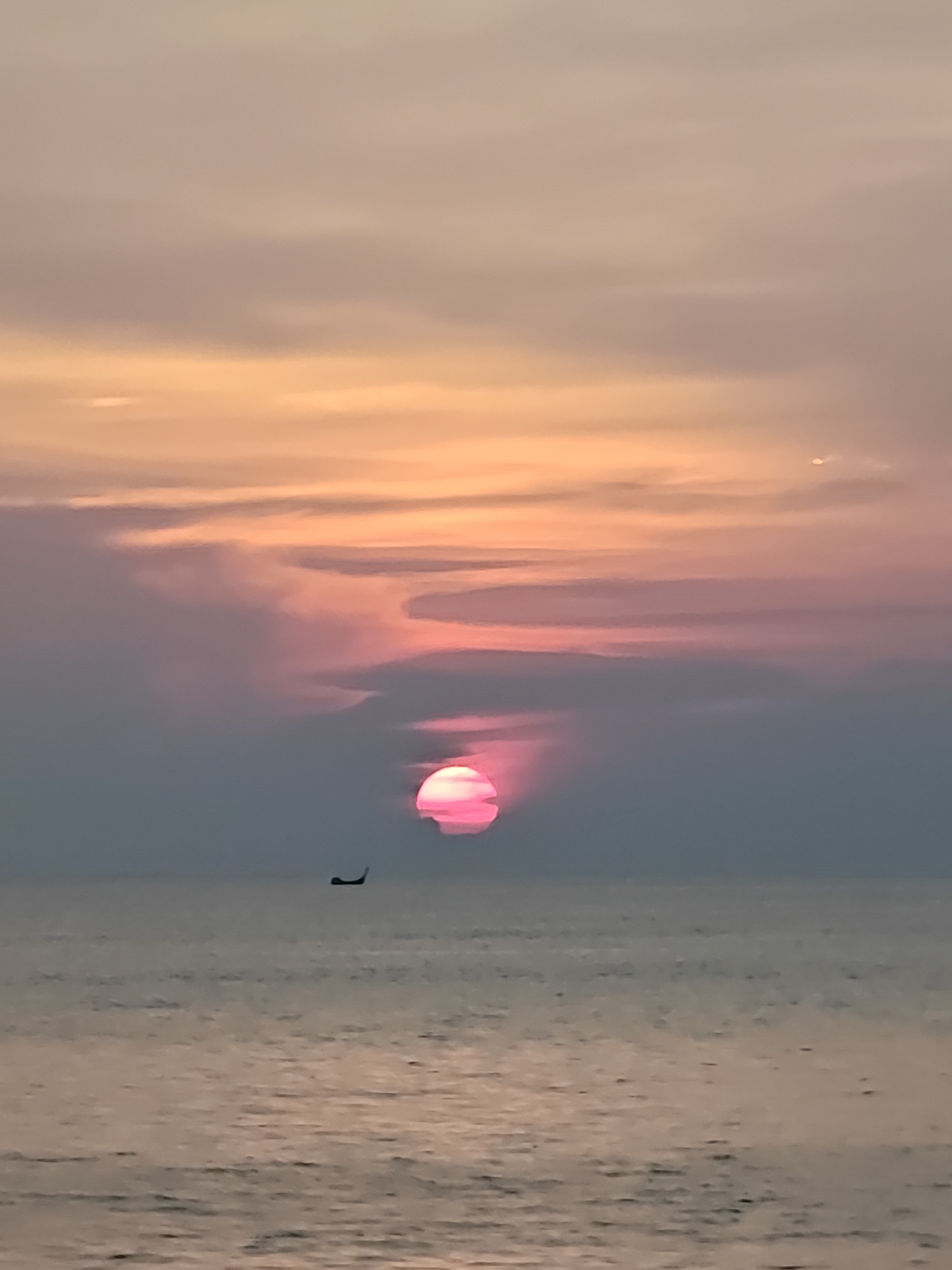 The image shows a sunset over the sea, with a pink sun partially obscured by clouds. A silhouette of a plane is visible above the water. The location is likely Lhokseumawe, Indonesia based on the prompt. The sky is a mix of orange, pink, and grey hues. The water reflects the colors of the sky.