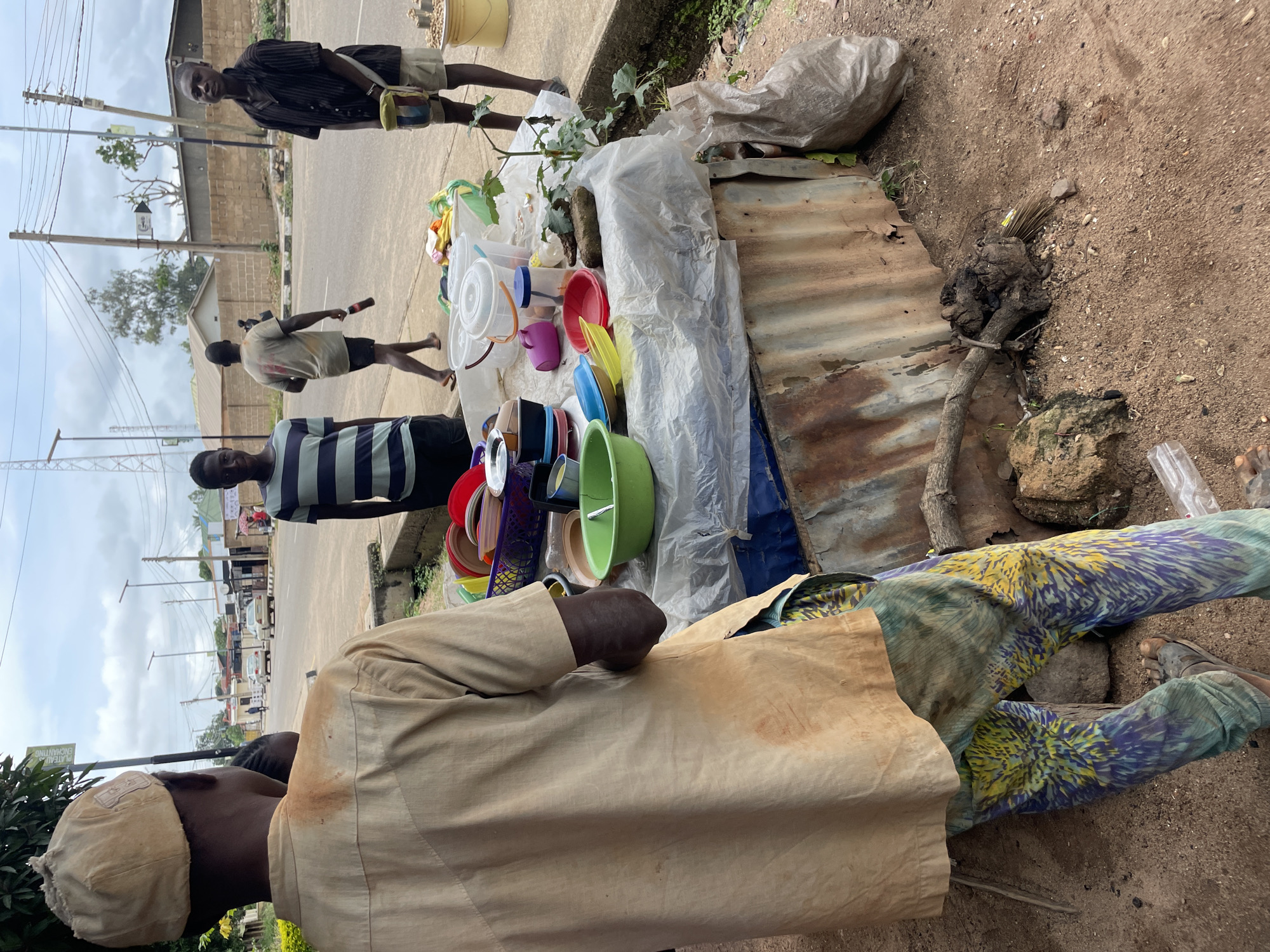 A street vendor in Jos, Nigeria, stands behind a table laden with various plastic household items and containers. The vendor, a young man wearing a light brown shirt and patterned trousers, is positioned in the foreground, with his back to the camera. To his right, a few other young men are visible, some standing by the roadside, others near the vendor's stall. The stall itself is covered with a white plastic sheet, displaying items like bowls, buckets, and plates in an assortment of colors. Behind the stall, a road with passing vehicles and buildings can be seen. The scene appears to be a typical day in a bustling urban environment.