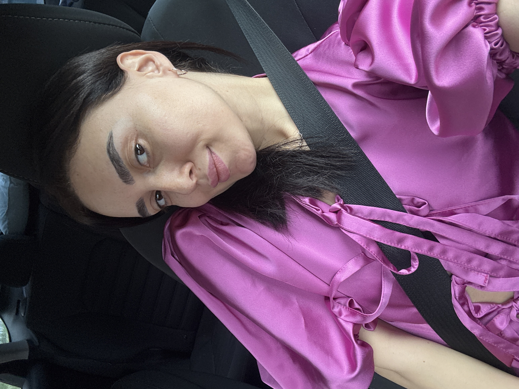 A woman with dark hair and fair skin is in the front passenger seat of a car, wearing a purple, satin-like top with puffy sleeves. She is wearing a seatbelt. The setting is indoors in a vehicle, possibly in transit. The city of Tbilisi, Georgia is not discernible from the image.